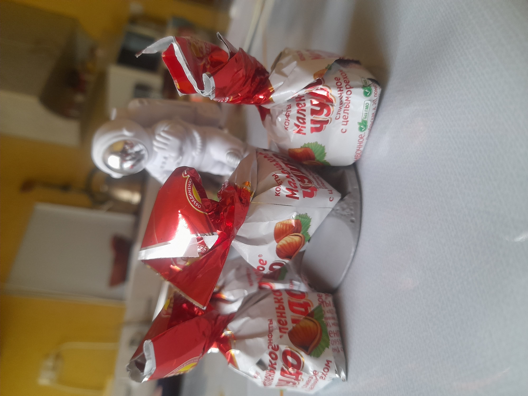 A close-up, eye-level shot captures three wrapped candies and a white astronaut figurine on a white, textured surface. The candies are individually wrapped in shiny red and white foil with images of hazelnuts and text in a foreign language, suggesting they are a confectionery product. The astronaut figurine, located behind the candies, is white with a reflective silver helmet. The background is blurred, showing hints of a kitchen environment with yellow walls and appliances. The overall scene appears to be indoors, likely in a domestic setting. There is no discernible indication of the city of Salsk, Russia, or any specific location beyond a general indoor setting.