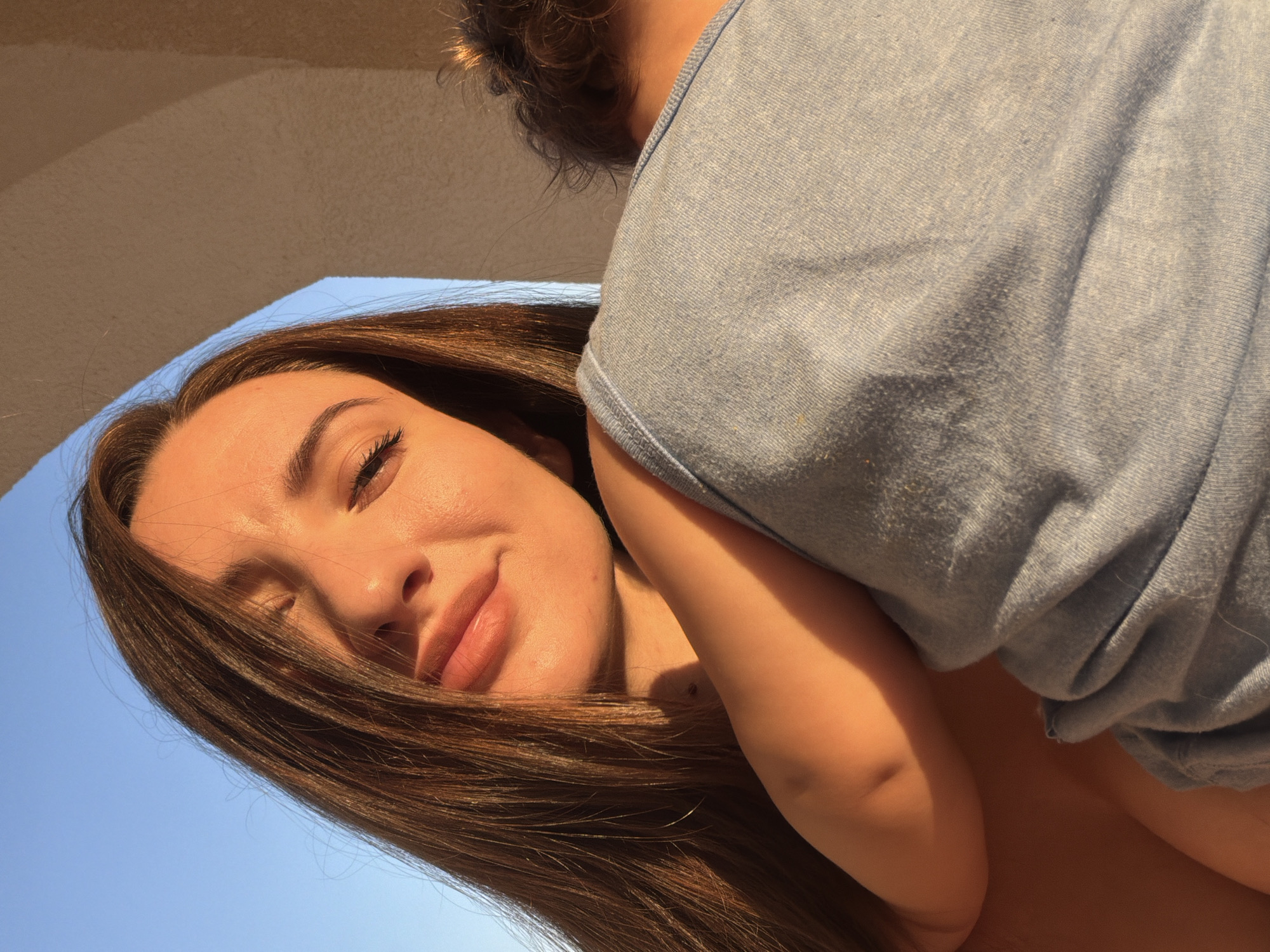 A young woman with long, dark brown hair and fair skin is shown in a close-up portrait. She is looking towards the viewer with her eyes partially closed, illuminated by warm sunlight. Her mouth is slightly open, and she appears to be smiling or about to speak. She is embracing someone, with their shoulder and the back of their head visible in the foreground, wearing a light blue t-shirt. The background shows a clear blue sky and an architectural element that appears to be a curved beige archway or overhang. The scene suggests a warm, outdoor setting, possibly during golden hour. There are no discernible elements that specifically place this image in Sharm El Sheikh, Egypt, beyond the potential for such a setting to exist there.