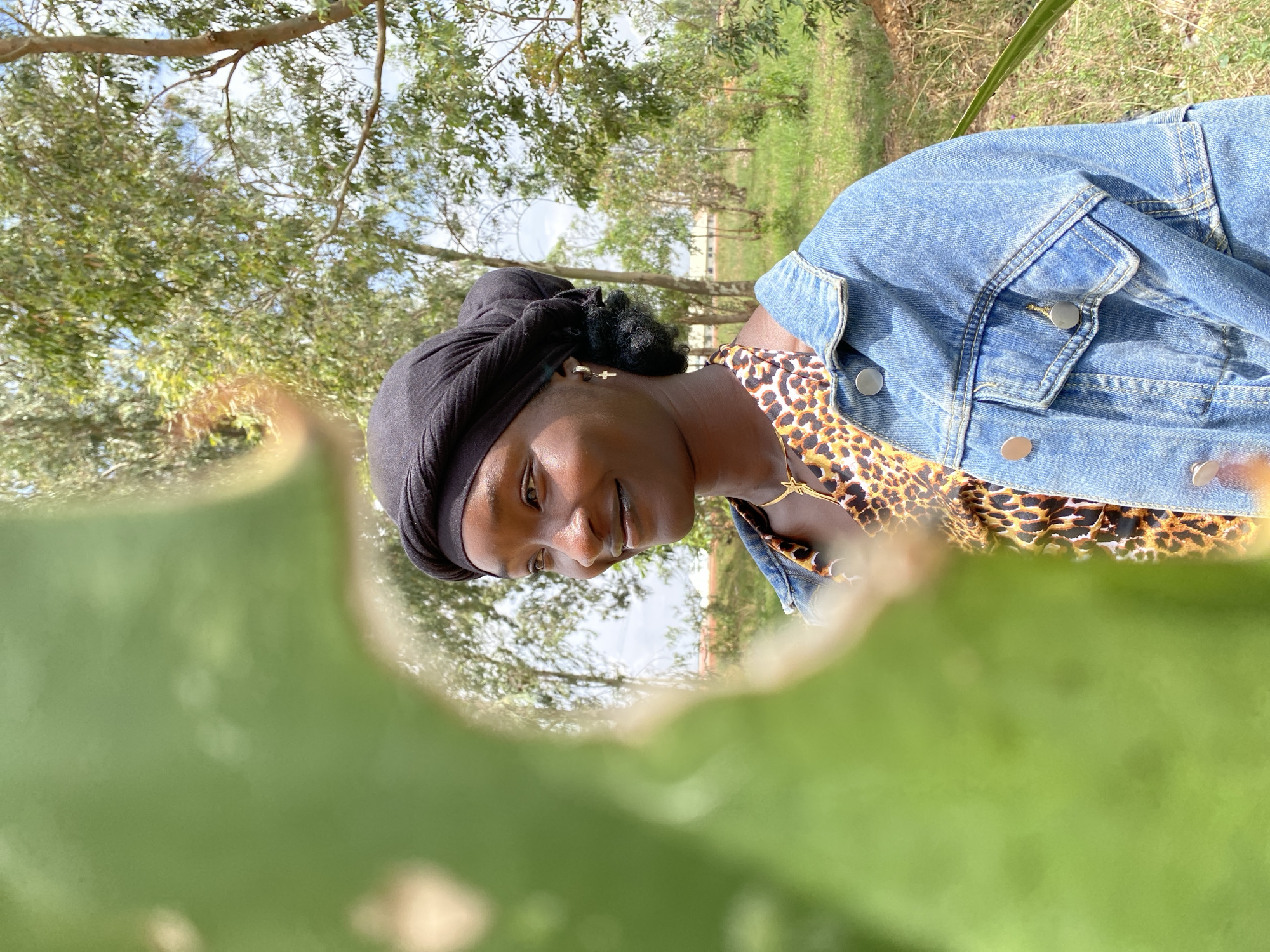 A young woman with dark skin and a black headscarf is depicted in the photograph. She is wearing a leopard-print top under a denim jacket. Her face is partially visible through a large green leaf in the foreground. The background features green grass and trees under a bright, slightly overcast sky. The specific location within Durimi, Nigeria cannot be determined from the visual information.