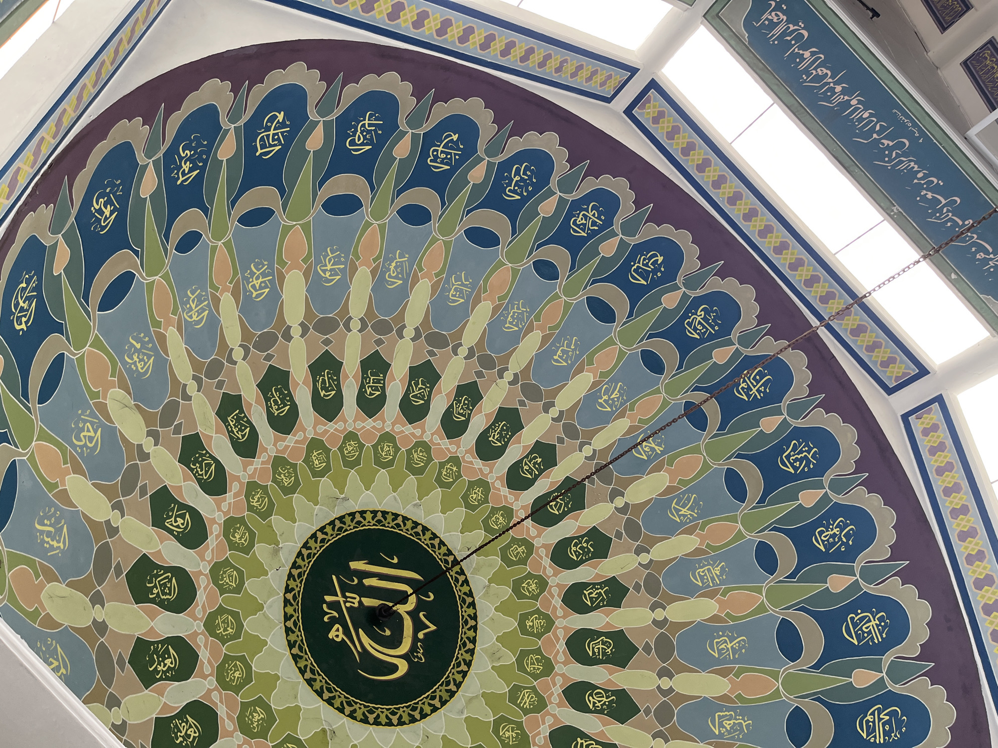 This media file depicts the interior of a mosque, specifically focusing on its ornate domed ceiling. The ceiling features a vibrant geometric pattern in various shades of blue, green, beige, and purple, with intricate Arabic calligraphy adorning sections of the design. The calligraphy is rendered in a bright yellow hue, making it stand out against the colorful background. In the center of the dome, a circular motif with similar yellow calligraphy is prominently displayed. A chain hangs down from the ceiling, suggesting the presence of a light fixture or decorative element. The overall impression is one of grandeur and religious artistry. The text visible in the image, as read by OCR, includes "الله" (Allah), along with several other Arabic words that appear to be divine names or phrases, such as "الشكون", "الحي", "السلط", "الظاهر", "الواسع", "المقدم", "القارب", "النهائي", "الصد", "الجليل", "الغياب", "واجد", and "النافقون ايه ؟ لنگ هم اخر". The location context provided is West Jakarta, Indonesia, which implies this is a mosque within that region. The image is taken from a low angle, looking up at the ceiling, and the lighting suggests it is daytime, possibly with natural light filtering in from skylights or windows integrated into the ceiling structure. No people are visible in the frame.