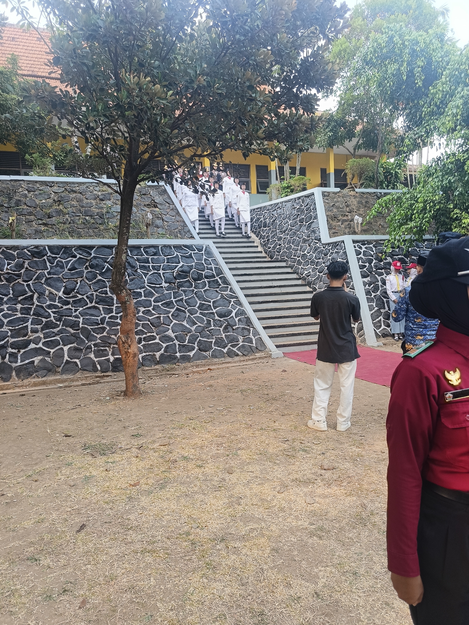 In Pengkol, Indonesia, a group of uniformed students in white attire ascend a stone-faced staircase leading to a yellow building. A lone tree stands to the left of the stairs, its bark textured and a light brown. In the foreground, a person in a dark shirt and light pants faces away from the camera, holding a phone. To the right, another person, partially visible, wears a red uniform with gold epaulets. The ground is a mix of dry grass and dirt, with scattered leaves. The sky is overcast.