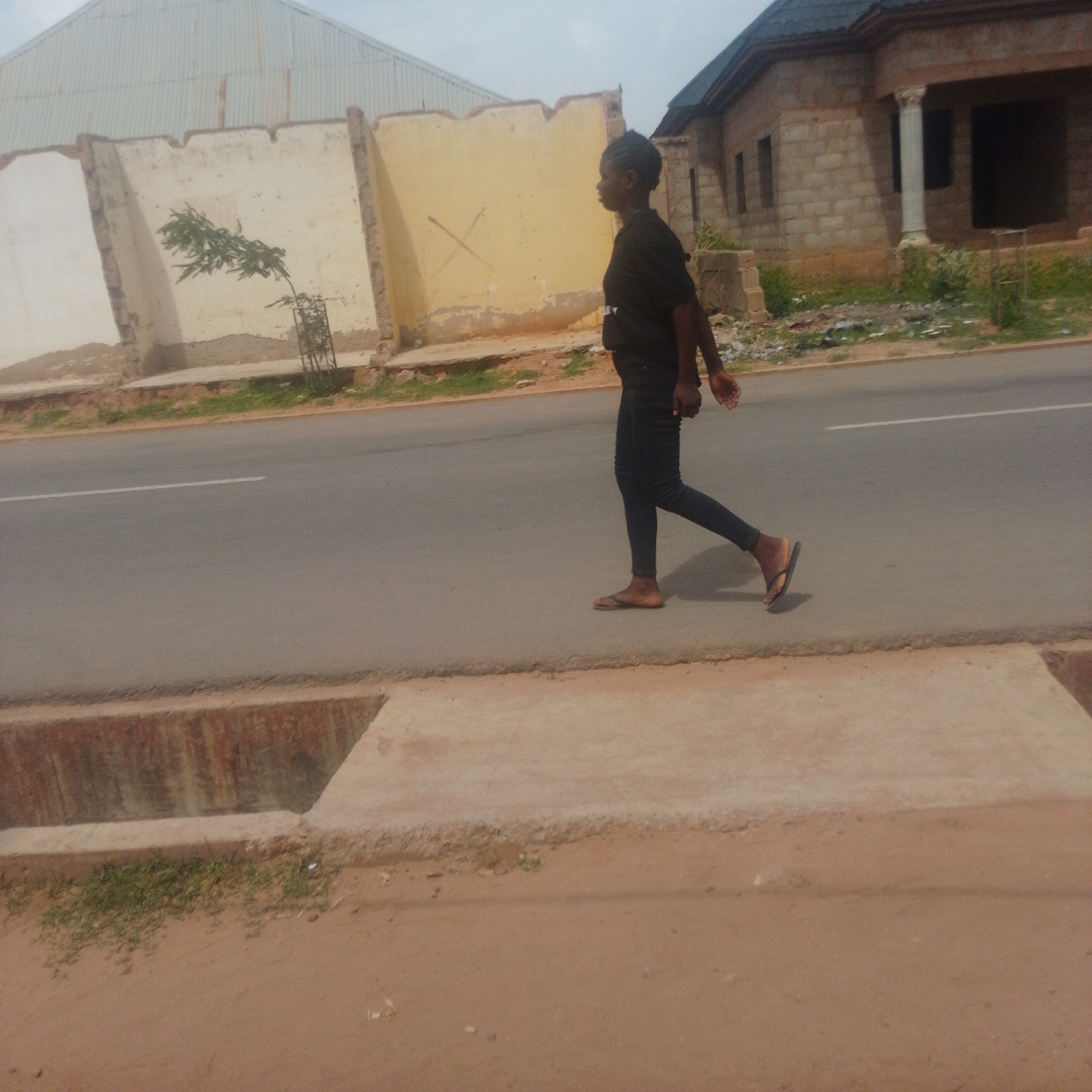 A young Black woman walks along a paved road in Gombe, Nigeria. She is wearing a black t-shirt and dark leggings, and appears to be wearing flip-flops. The woman is walking from left to right in the frame, and her body is in profile, facing towards the right side of the image. Her arms are bent at the elbows, and her right arm is extended slightly forward, as if in mid-stride.
The background of the image shows a developing urban or semi-urban environment. To the left of the woman, there is a partially constructed building with a corrugated metal roof. Adjacent to this building is a wall, half-demolished and painted in muted colors. A small, leafy tree grows near the wall, protected by a metal cage. Further to the right, a brick house is under construction, with columns framing an entrance. Debris and vegetation are scattered around the construction sites, suggesting an ongoing development or an area in transition.
The road itself is asphalt, with a white line marking the lane. A concrete gutter runs along the side of the road closest to the viewer. The ground on the viewer's side of the gutter is sandy with some sparse grass. The sky is overcast, indicating a cloudy day, and the lighting is diffused, suggesting it might be morning or late afternoon. There are no discernible emotions visible on the woman's face, and the overall impression is one of everyday activity in a Nigerian setting. No visible text is present in the image.