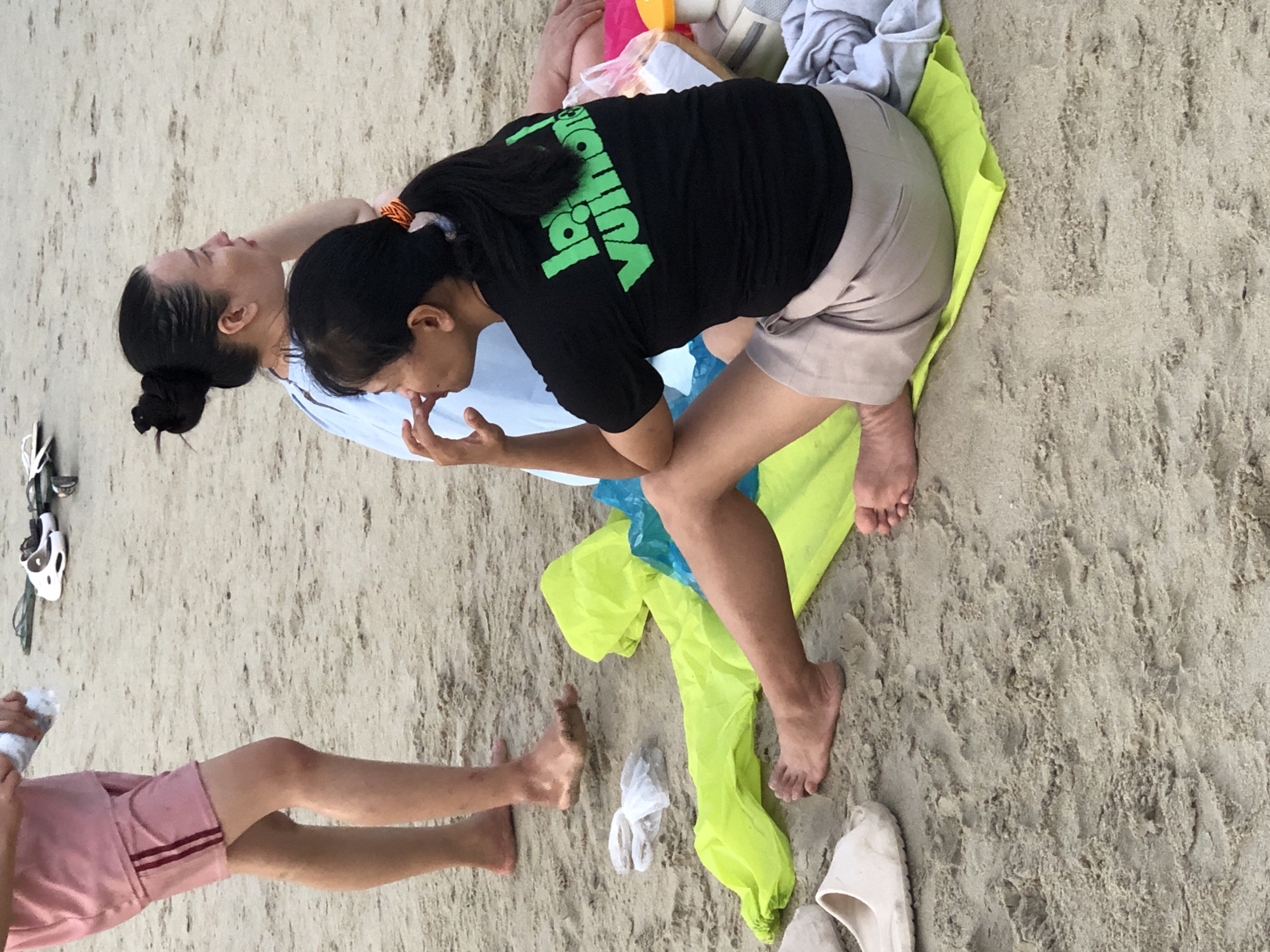 The image shows a beach scene in Đà Nẵng, Vietnam. In the foreground, two women are sitting on the sand. One woman is wearing a black t-shirt with green text and a light-colored skirt. The other woman is wearing a light blue top. Part of a person's legs, wearing pink shorts and a pink shirt, are visible on the left side of the image. Several objects are scattered on the sand, including a bright yellow towel or blanket, some white plastic bags, and a pair of white sandals. In the background, more beachgoers and some footwear can be seen. The sand appears light brown and textured, typical of a beach environment.