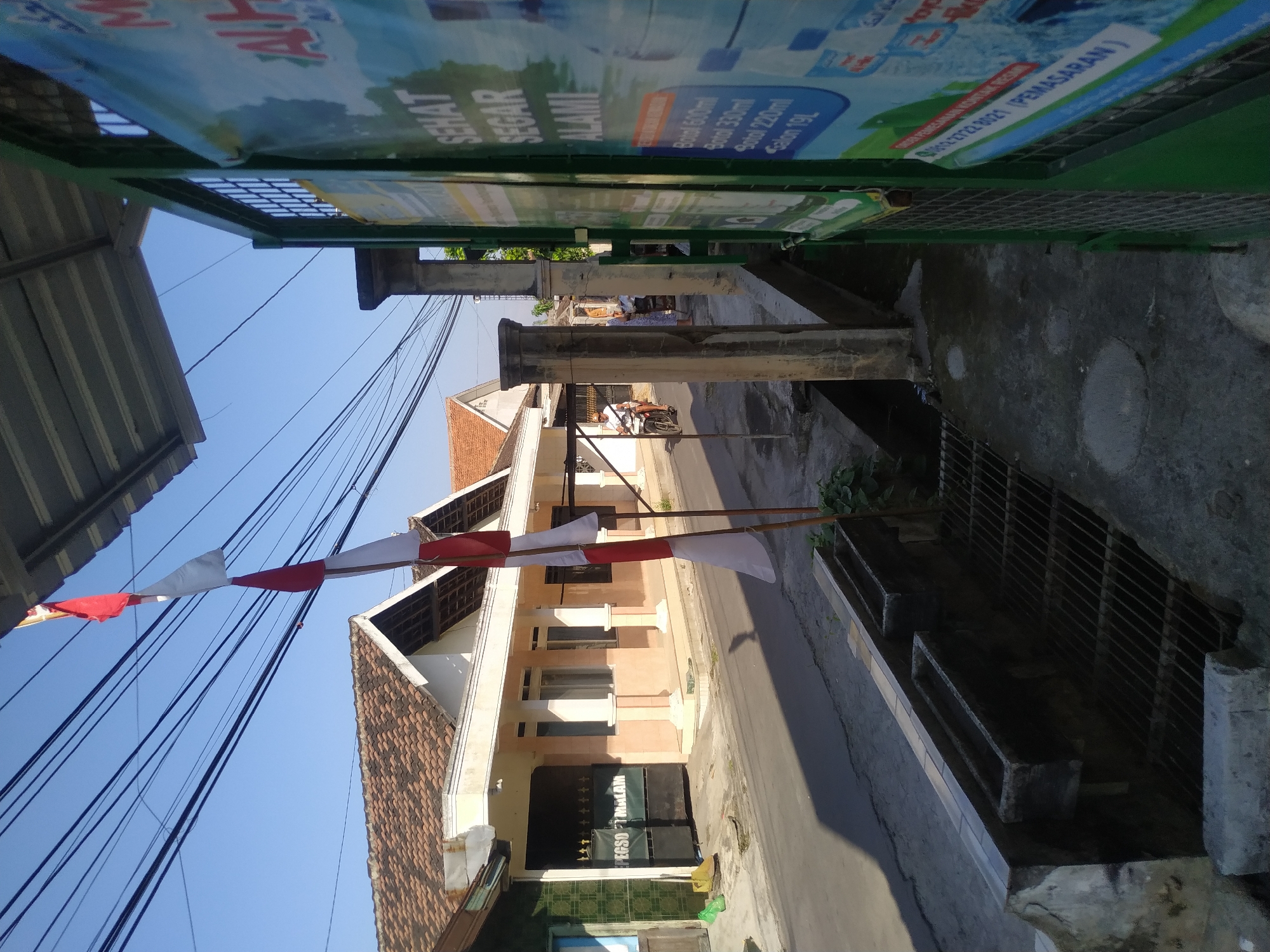 The image shows a street in Batan, Indonesia. The scene is captured in daylight with clear skies. In the foreground, a drainage grate and a concrete channel are visible. To the right, a green metal fence surrounds a property, with a large advertisement poster partially visible. The poster features text in Indonesian, including "SEHAT SEGAR ALAMI" (Healthy Fresh Natural) and lists bottle sizes for what appears to be water or a beverage. Further into the scene, a red and white Indonesian flag is displayed on a pole. On the left side of the street, a row of buildings with tiled roofs and beige facades line the road. One building has a dark-colored gate with gold text reading "PEGSOF MALAM". A person on a motorcycle is riding down the street, and another person is standing near the buildings. Electrical wires are strung across the sky above the street.
