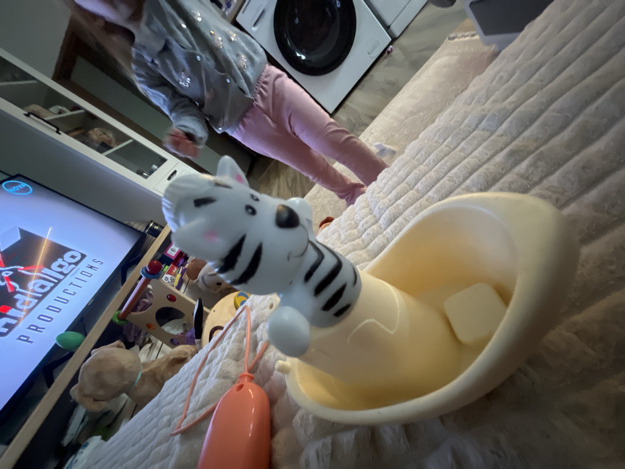 A young child is standing in an indoor setting with a washing machine in the background. In the foreground, a zebra-striped toy is positioned in a yellow container. A television displaying "Hidalgo Productions" can be seen on the left, along with other toys in the periphery. The setting appears to be a home environment.