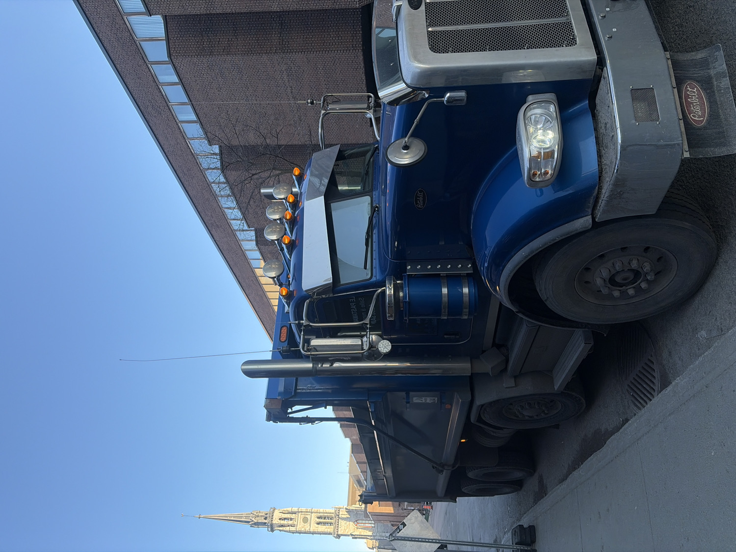 The image features a large blue Peterbilt semi-truck, specifically the front and driver's side of the tractor unit and part of its dump trailer, parked on a city street in Montreal, Canada. The truck is the primary subject, displaying a prominent chrome grille, multiple amber marker lights along the roofline, large chrome exhaust stacks, and a chrome fuel tank. The bright blue paint of the cab is reflective. "Peterbilt" is visible on the front fender/mudflap and as a logo on the cab door.
In the background, a multi-story building with red brick walls and horizontal strips of windows runs along the upper part of the frame. The sky is a clear, bright blue, indicating a sunny day. In the distance, towards the lower left of the frame, a tall and intricately designed church steeple, characteristic of Gothic revival architecture, rises against the blue sky. The ground beneath the truck consists of gray pavement, with a visible drain cover near the front wheel. The truck appears to be stationary. The overall scene suggests a typical urban setting during daytime.