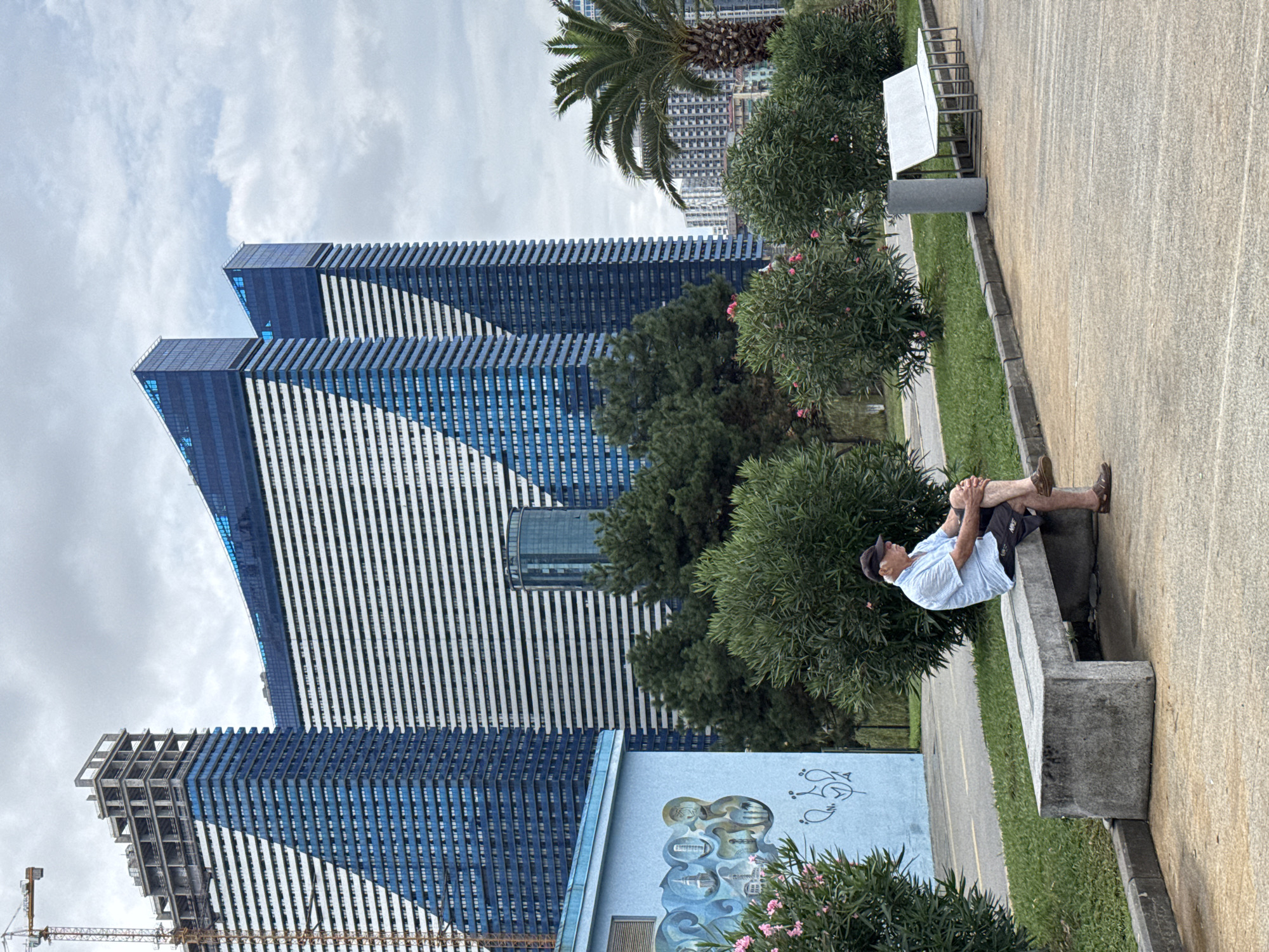 An elderly Caucasian male wearing a light blue short-sleeved collared shirt, dark shorts, a dark baseball cap, and brown slip-on shoes is sitting on a concrete bench. He is positioned on the right side of the bench, with his legs crossed at the ankles and his hands clasped on his right knee. He is looking to his right, away from the camera. The bench is made of concrete and is situated on a paved walkway. To the left of the man is a light blue building with a mural depicting stylized buildings and birds. Behind the man and the bench, there are lush green bushes and trees. In the background, three distinctive modern skyscrapers with blue glass facades and white horizontal stripes dominate the skyline. A construction crane is visible to the far left, attached to one of the incomplete skyscrapers. The sky is overcast with scattered clouds. The scene is set in Batumi, Georgia, likely on a promenade or public space near the Black Sea coast.