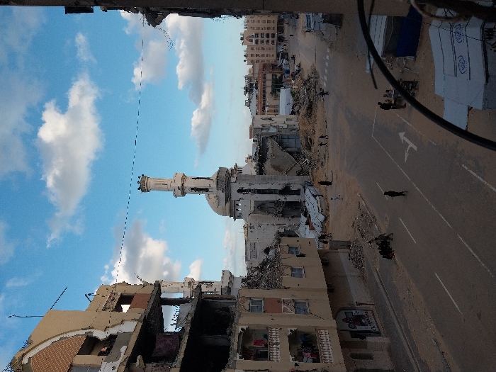 This image depicts a wide street in a severely damaged urban environment in Khan Yunis, Palestinian Territory, under a sky with scattered white clouds. In the mid-ground, the prominent structure of a mosque stands heavily damaged, featuring a partially collapsed white dome and a tall, slender minaret with a distinctive upper section. The mosque's main body is largely reduced to rubble, with structural elements visible.
On the left side of the street, a multi-story residential or commercial building exhibits extensive destruction, with large sections of its facade missing, revealing interior spaces and exposed concrete and rebar. Clothes are hanging on a lower-level balcony of this building. Rubble and debris are accumulated at its base and extend into the street. A damaged building is partially visible on the far right foreground.
The paved road in the foreground and mid-ground is significantly covered with sand and scattered debris. A white directional arrow is painted on the road surface, pointing right. Approximately 15-20 individuals are present in the scene; several are walking singly across the road or amidst the rubble, while a small group is gathered near the base of the damaged building on the left. Other figures are observable near the mosque's ruins and further down the street. In the lower right foreground, a portion of a makeshift shelter or tent, possibly a humanitarian structure with visible text, is present. Overhead utility lines traverse the upper portion of the frame.