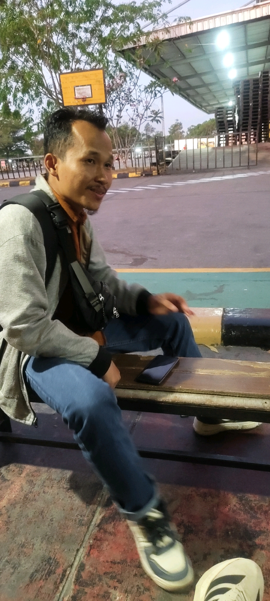 In Nusa Indah, Indonesia, a man is seated on a wooden bench outdoors. He is wearing a grey jacket over an orange shirt and blue jeans, with a black bag slung across his chest. A smartphone lies on the bench next to him. The background reveals a fenced area with a covered structure and stacks of black pallets. A yellow basketball hoop is visible in the distance, amidst trees. The ground is a weathered surface with painted lines.
