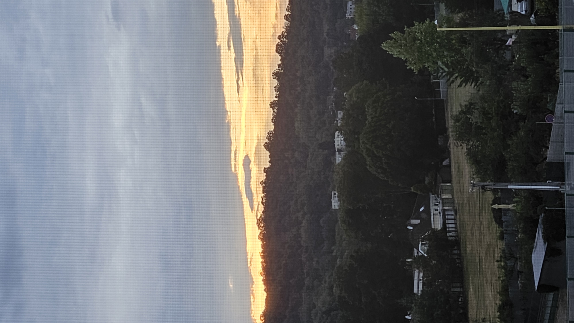 The image depicts a hazy, underexposed view of a landscape at dusk or dawn, possibly from a window. The sky is a pale blue with diffused light, suggesting clouds or atmospheric conditions. A sliver of bright orange light is visible near the horizon, behind a silhouetted line of trees and vegetation that form a hilly terrain. Below the hill, several buildings are faintly discernible amongst the trees, appearing to be residential structures. The foreground shows more dense vegetation and what appear to be fences and a small structure, possibly a shed. The overall impression is a somewhat obscured and distant view of a semi-rural or suburban area. The location is stated to be Prešov, Slovakia.