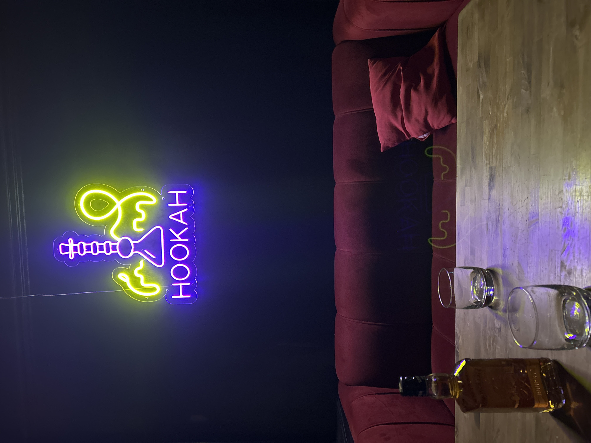 A dim interior shot featuring a red velvet sofa with a matching pillow, a wooden table with two empty glasses and a partially visible whiskey bottle, and a neon sign displaying "HOOKAH" in yellow and purple. The background wall is dark, contributing to the overall low-light ambiance. The location in the city of Klin, Russia, cannot be confirmed from this image.