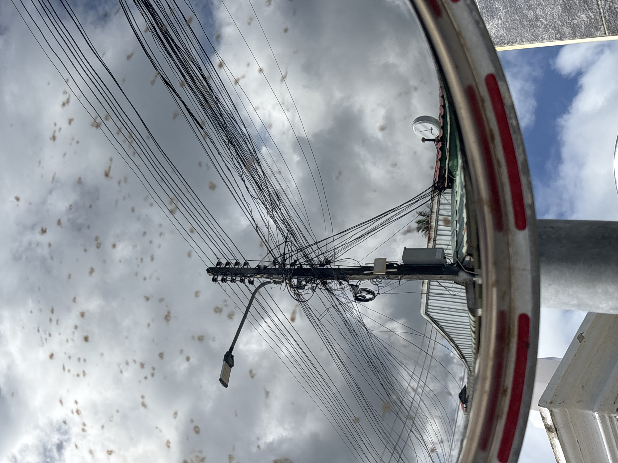 The image shows a street scene, likely in Rawai, Thailand, as indicated by the style of the utility pole and overhead wiring, commonly found in tropical urban environments. The primary focus is a wooden utility pole laden with a dense network of black electrical and communication wires. A street lamp is attached to an arm extending from the pole. Below the wires and to the right of the pole, a white box is mounted, possibly an electrical junction or communication equipment. In the background, parts of a corrugated metal roof and another building with a tiled green roof are visible, suggesting a commercial or residential area. A round, white clock with black hands is mounted on the green roof. The sky is cloudy with some patches of blue sky peeking through. The foreground is dominated by a curved, metallic barrier with red reflective strips, suggesting a traffic mirror or a protective barrier. The mirror aspect of the foreground indicates that this photo may have been taken with a wide-angle or convex lens, possibly from a traffic mirror itself, which would explain the distorted reflections of the sky and buildings. There are no people visible in the image. The overall scene depicts a typical roadside environment with a complex web of infrastructure.