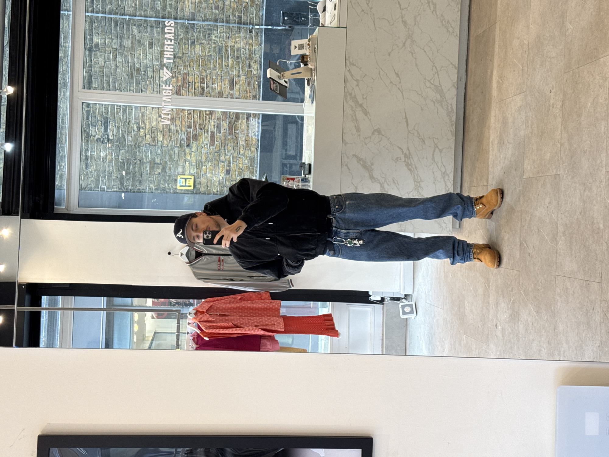 A person wearing a black hoodie, blue jeans, and tan boots is standing in front of a mirror, taking a selfie with a smartphone. The mirror reflects clothing on hangers, including a gray jacket and a set of reddish-orange clothing. Visible through the window behind the person is a brick building, a street, and the storefront of "Vintage Threads". The storefront and building design indicate this is in the city of London, United Kingdom. The floor and the wall next to the window are light colored and of marble.