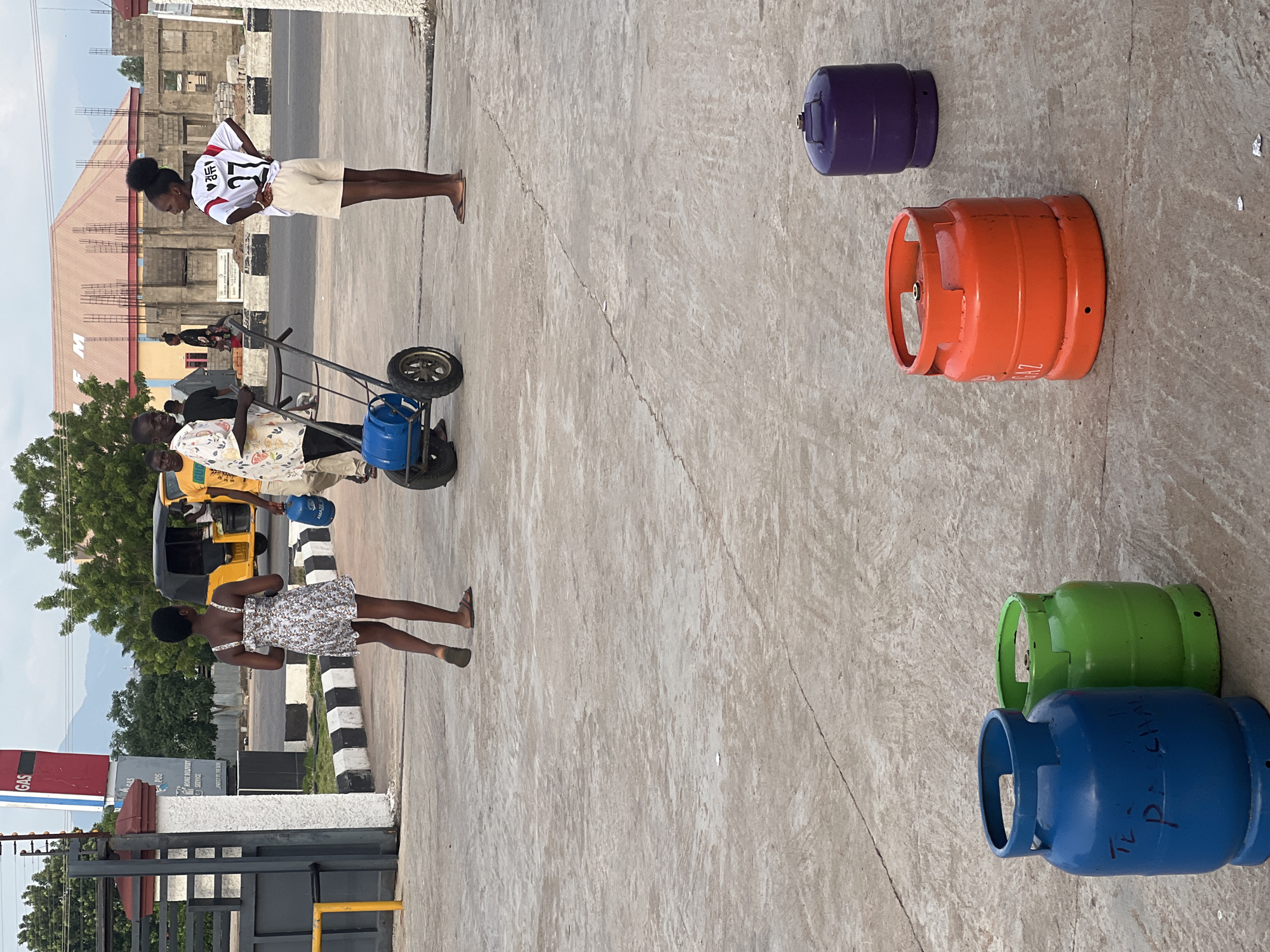 In the city of Jalingo, Nigeria, a gas station forecourt is depicted with several people and gas cylinders. In the foreground, a blue, a green, and an orange gas cylinder stand on the paved ground. A purple gas cylinder is visible further back. In the middle ground, a young woman in a short floral dress and sandals walks away from the camera. Beside her, a man in a white shirt with a colorful pattern pushes a cart carrying a blue gas cylinder. Another man in a yellow shirt is partially visible behind him. To the right, a young woman in a white and red sports jersey and shorts stands with her hands on her hips, facing the cart. In the background, a yellow auto-rickshaw is parked, and the facade of a building under construction is visible, along with a "GAS" sign on a red and white structure. The ground is a worn concrete surface.