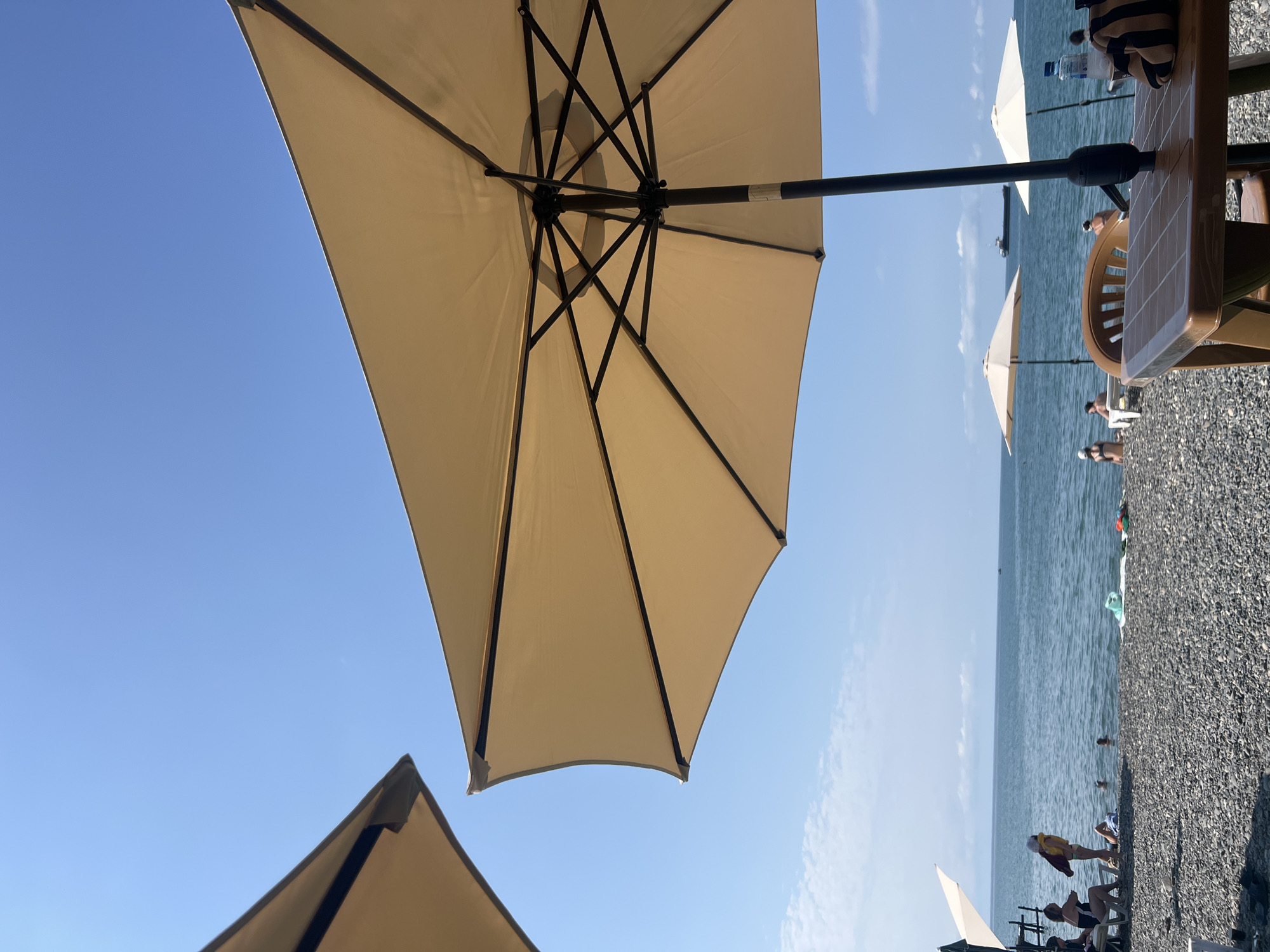The image depicts a sunny day at a pebble beach in Sokhumi, Abkhazia. The foreground is dominated by the underside of a large, beige beach umbrella, its black metal frame clearly visible. To the left, another beach umbrella's corner is visible against the clear blue sky.
In the middle ground, the blue sea stretches to the horizon, where a cargo ship is visible. Several other beach umbrellas are set up on the beach, with some people seated beneath them. A few individuals are in the shallow water. The beach itself is covered in dark grey pebbles.
In the far background, a faint hazy line suggests distant land. The overall atmosphere is one of leisure and relaxation on a warm day at the seaside.