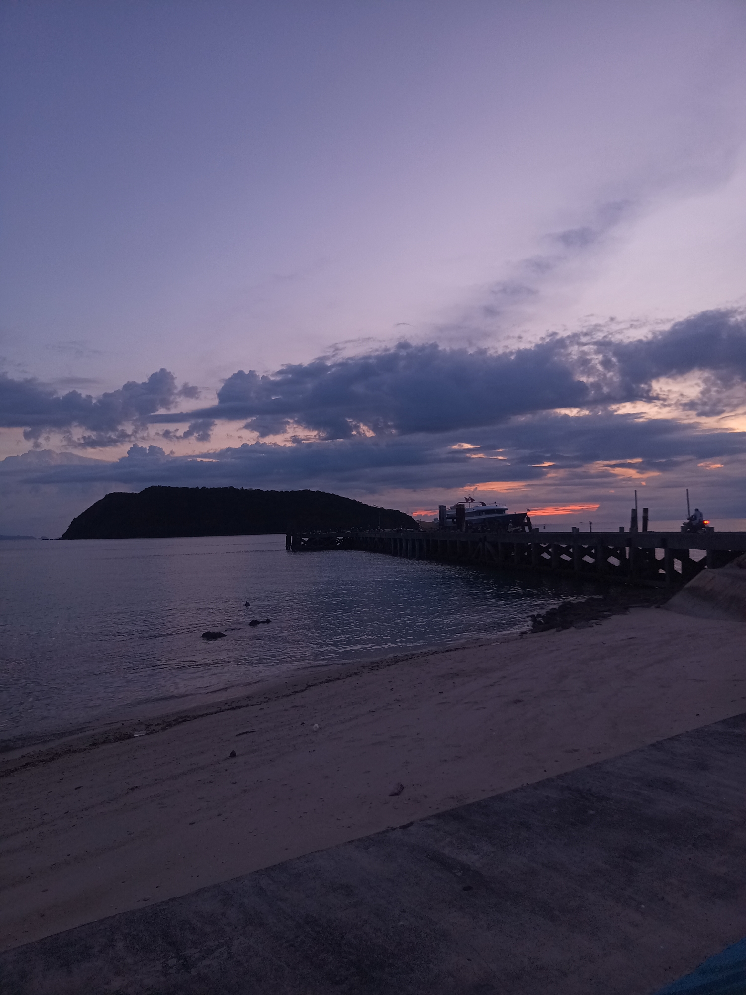A wide shot captures a serene coastal scene at dusk, likely in Koh Phangan, Thailand. The sky is a canvas of soft purples and grays, with scattered clouds tinged by the fading sunlight. A dark, mountainous island dominates the left side of the frame, silhouetted against the horizon. In the foreground, a sandy beach transitions to a concrete embankment.
Extending from the right side of the image is a weathered wooden pier. A ferry or large boat is docked at the pier, its structure partially obscured by the low light. A few people are visible on the pier, one appearing to be riding a scooter, its headlight casting a faint orange glow. The water is calm, reflecting the muted colors of the sky. The overall mood is tranquil and peaceful, indicative of the twilight hour. No text is visible in the image.