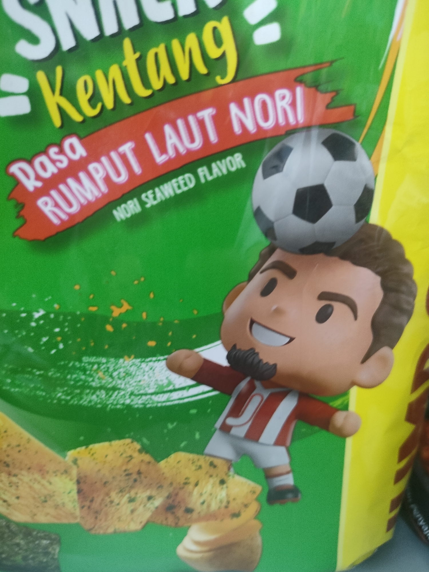 The image is a close-up shot of a bag of "Kentang Rasa Rumput Laut Nori" (Potato Seaweed Nori Flavor) potato chips, featuring a cartoon caricature of a soccer player balancing a soccer ball on his head. The bag is predominantly green with yellow and red accents, and the text is in Indonesian. The caricature is dressed in a red and white striped soccer jersey and white shorts, with black soccer shoes. The soccer ball is grey and black. In the foreground, several potato chips are visible, scattered across the bottom of the image. The lighting is bright and appears to be from a flash, with some glare on the packaging. The background is a plain yellow surface, likely the side of the bag or a display. The image appears to be taken indoors, possibly in a store or at home. There is no information in the image to indicate its location in Desa Banyumas, Indonesia.