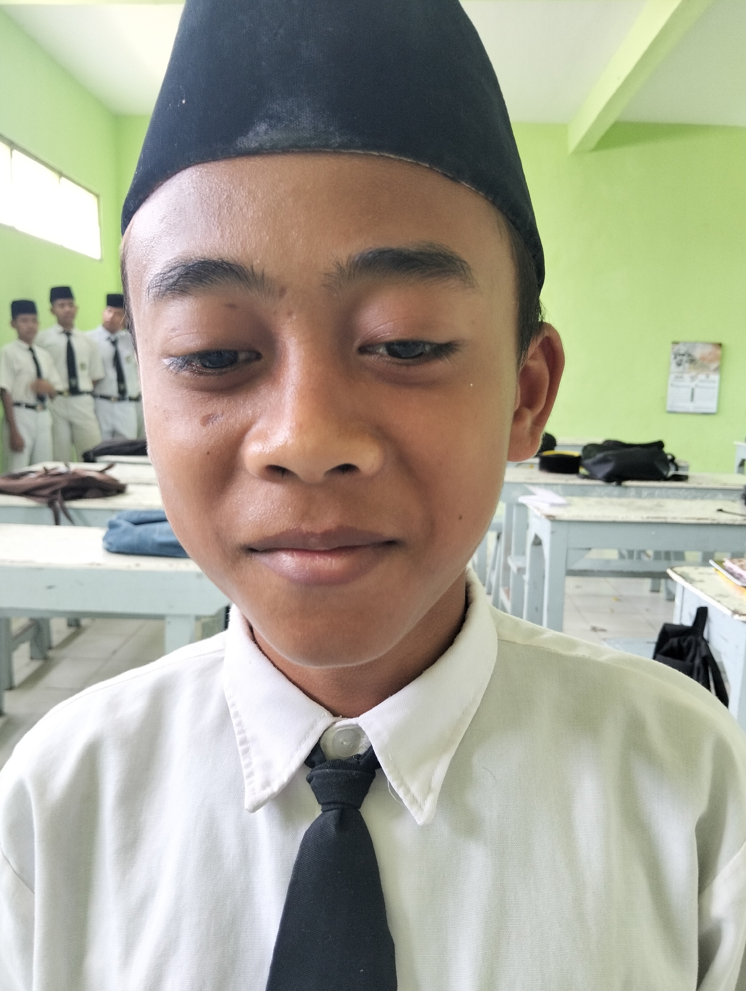 A close-up, frontal view shows a young boy wearing a black peci hat and a white collared shirt with a black tie. He is looking directly at the camera with a neutral expression. The background, out of focus, reveals a classroom environment with several other boys in similar attire standing in the distance. Desks and chairs are visible, along with pale green walls and a window. The overall scene suggests a school setting in Kranji, Indonesia.