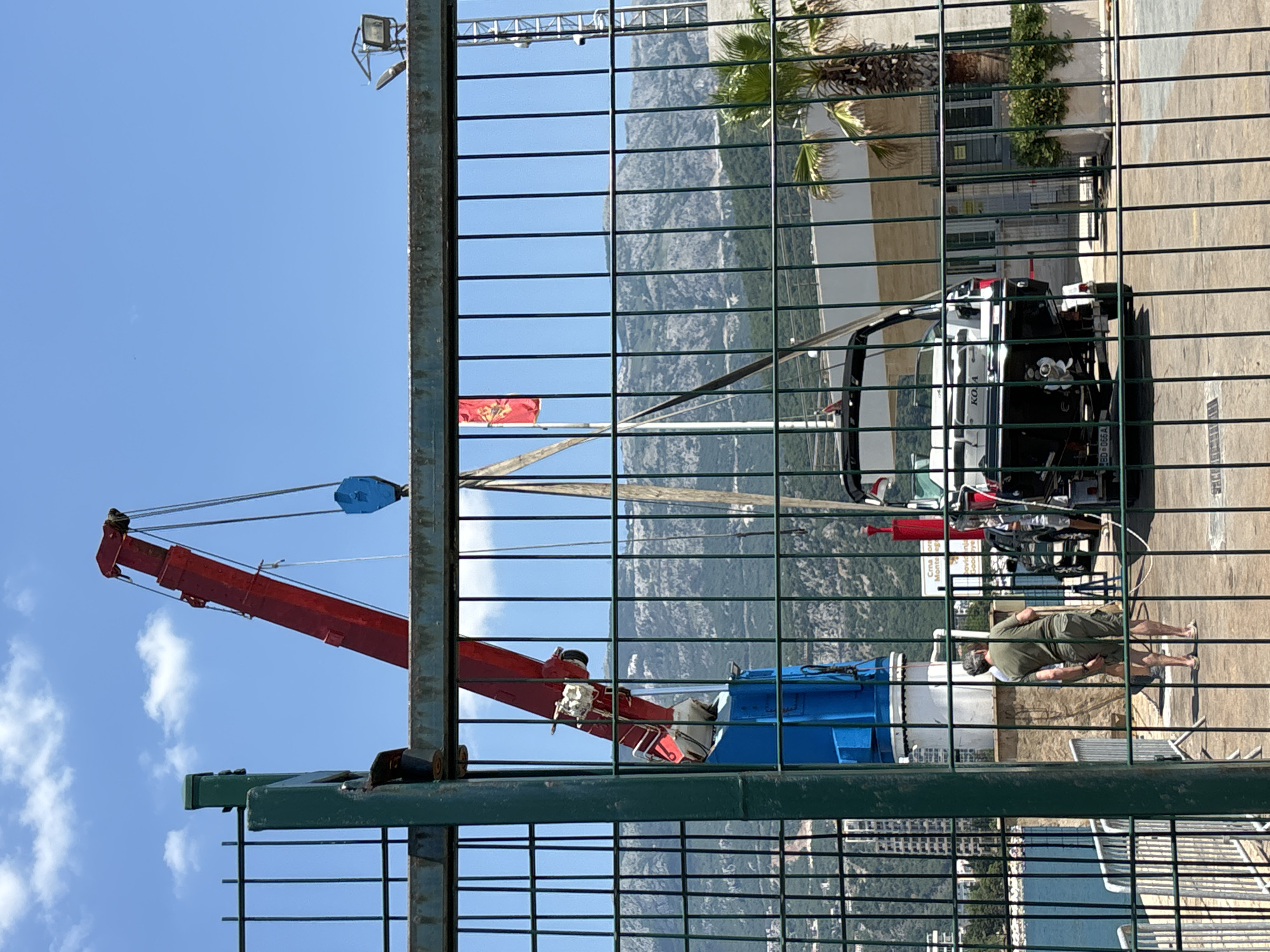 In Budva, Montenegro, a red crane is actively lifting a black boat from the water. The boat is on a trailer and is being maneuvered over a paved area adjacent to a body of water. Two men are present; one, wearing a green t-shirt and shorts, is standing on the paved area near the crane's base, while another person, partially obscured by the fence, is near the boat. The scene is framed by a green metal fence with horizontal and vertical bars. In the background, there are buildings, lush greenery, palm trees, and a mountain range under a clear blue sky with scattered white clouds. A red flag with a yellow emblem is visible on a flagpole to the left of the crane.