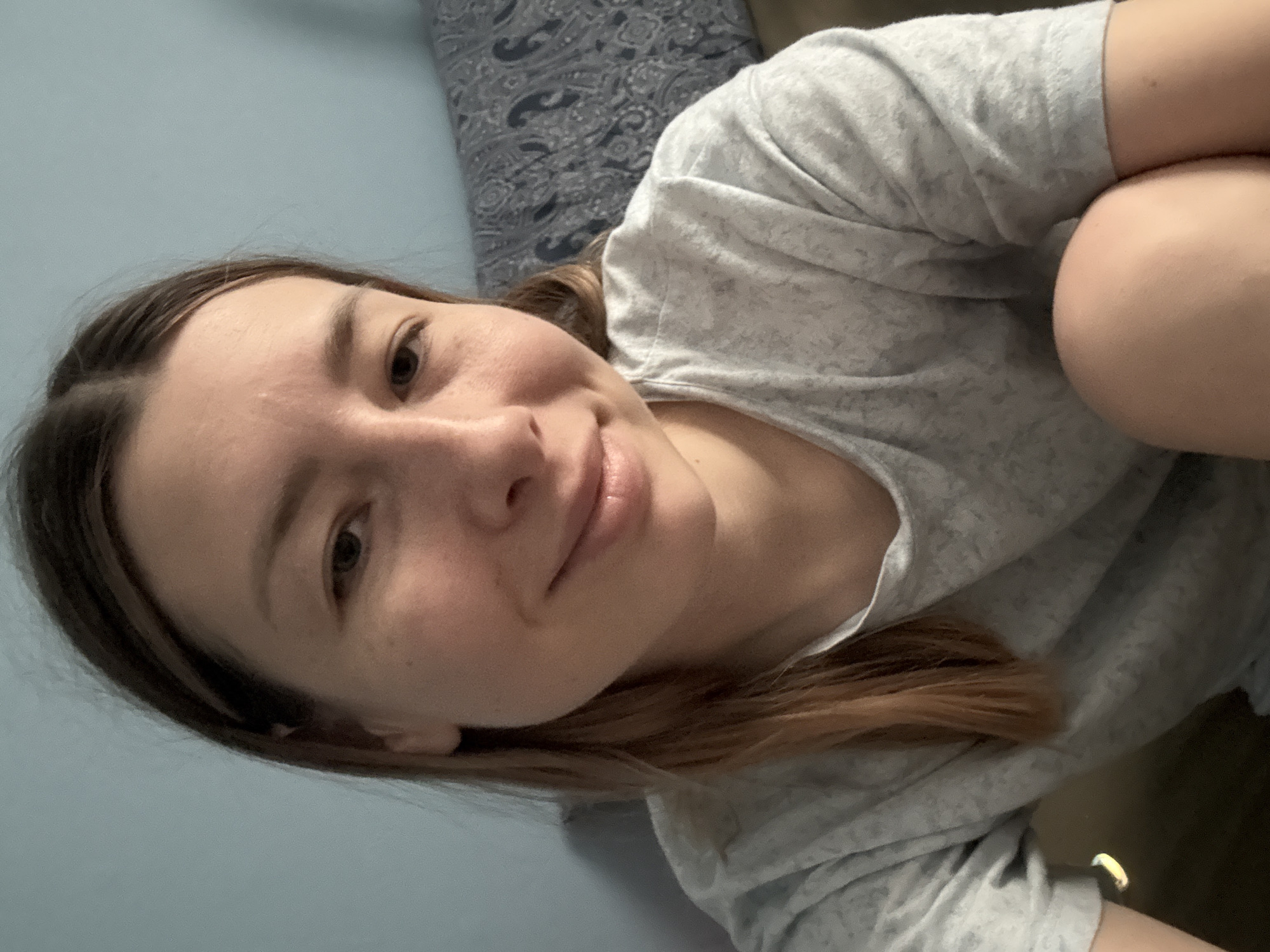 A woman with brown hair is lying down indoors. She is wearing a gray patterned t-shirt. The background includes a blue wall and a patterned pillow. The location in Moscow, Russia, cannot be determined from the image.