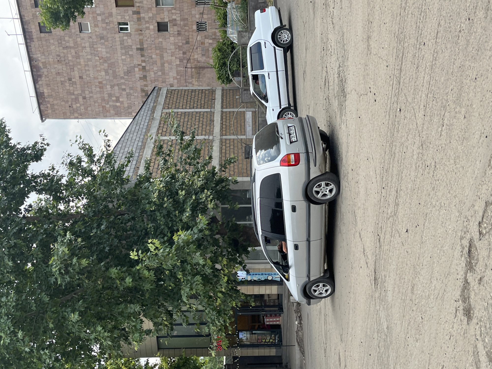 The image shows a street scene in Yeghegnadzor, Armenia. In the foreground, a silver minivan is parked on a dirt and gravel lot, facing left. The minivan has its rear facing the camera. A white sedan is parked to the right of the minivan, also facing left. The buildings in the background are made of brick and stone. A large tree with green leaves is in the left part of the image. The sky is overcast. There is no visible interaction between the people in the vehicles and any other individuals.