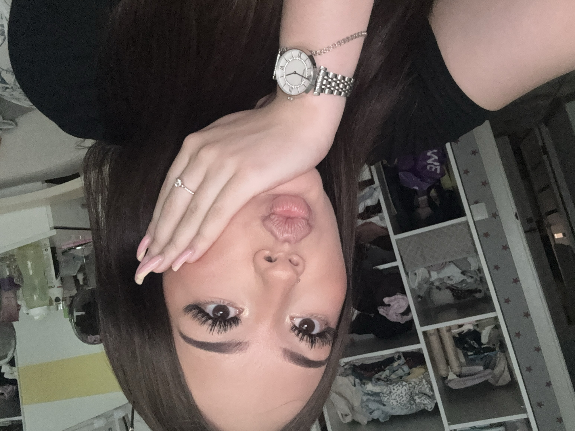 In a brightly lit room, a young woman with dark hair and long eyelashes poses for a selfie. Her hand is placed on her cheek, and her lips are puckered. She wears a black shirt, a silver watch with a white face, and a delicate ring on her right hand. Behind her, shelves are filled with clothing, personal items, and decorative stars on the wall. The environment suggests a personal living space, possibly a bedroom or dressing area, within a residential setting. The specific location in the city of Naberezhnye Chelny, Russia, cannot be determined from the visual information provided.