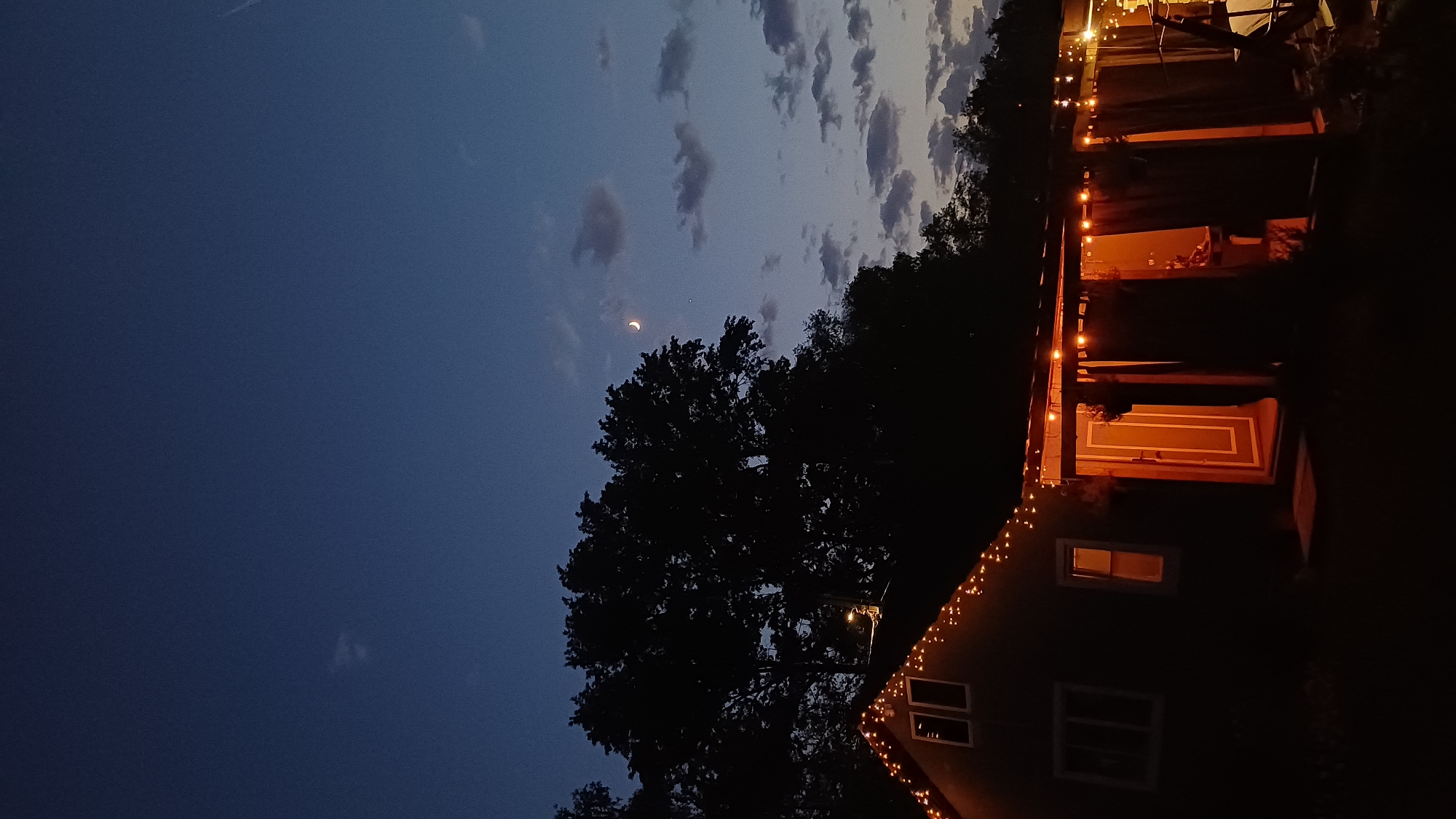 The image captures a night scene, likely in a rural area of Timokhovo, Russia. A dark silhouette of a large tree dominates the foreground, partially obscuring a two-story house on the right. String lights adorn the house's roofline and porch, providing illumination. The sky transitions from a dark blue to a lighter shade with scattered clouds, and a crescent moon is visible. The image suggests a calm, quiet evening setting. No people are visible.