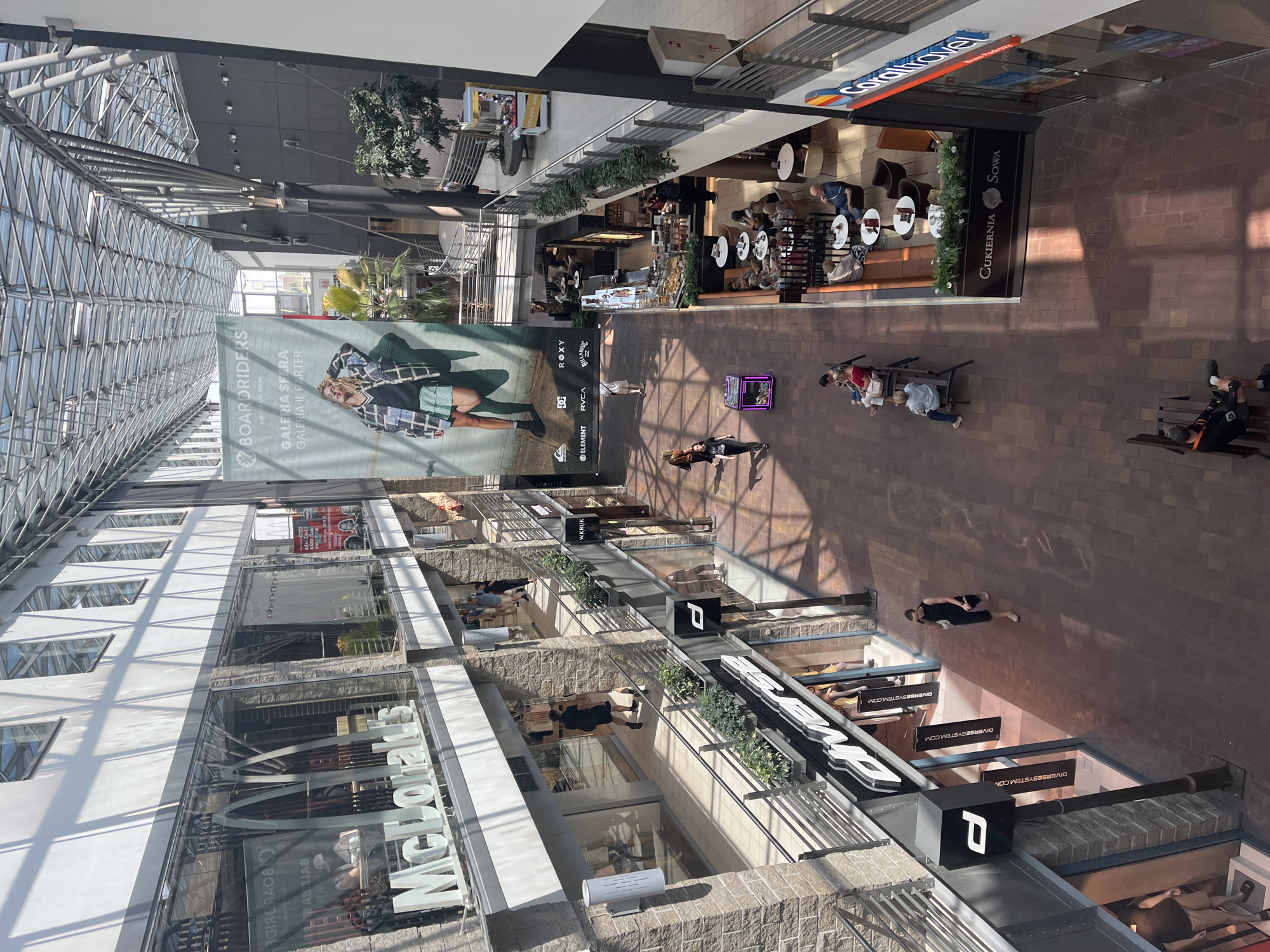 The image depicts an interior view of a multi-level shopping mall. Sunlight streams in from a glass ceiling, casting shadows on the polished floor. On the left, a McDonald's restaurant is visible with its prominent signage. Adjacent to it, there are other retail stores with glass storefronts, some displaying mannequins. On the right side, a cafe area with tables and chairs is occupied by patrons. Further down the corridor, a travel agency named "Coral Travel" is located. A large banner advertising "Boardriders" hangs from the ceiling, featuring a model in surf-inspired clothing. People are seen walking, sitting at cafes, and browsing in the shops. The architecture of the mall includes exposed metal beams and stone accents on the walls.