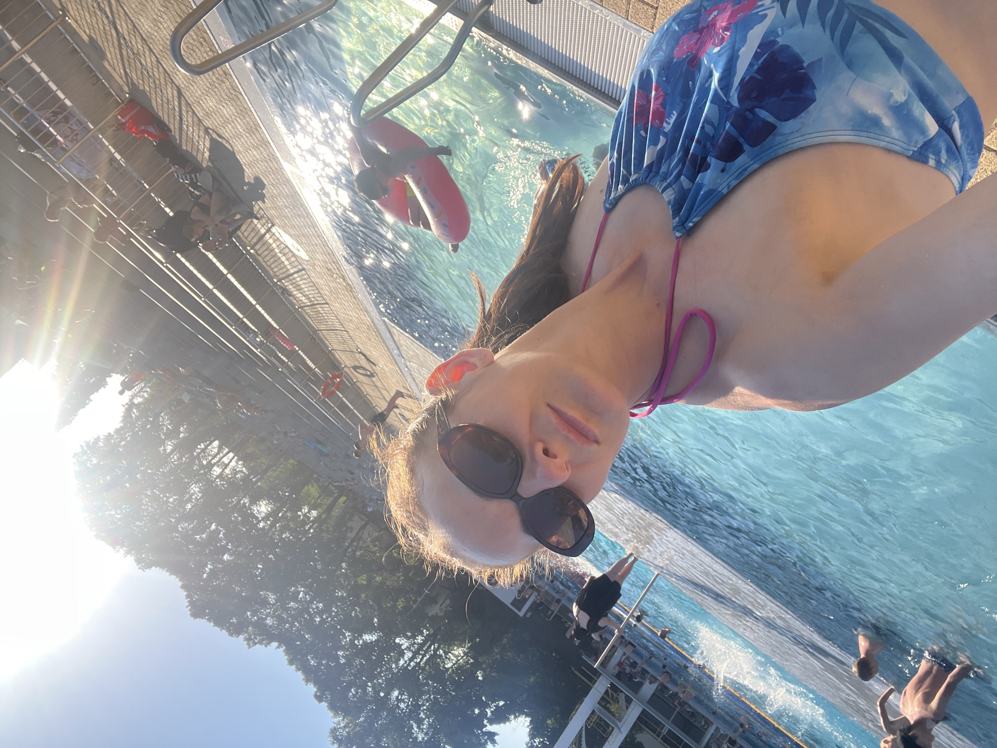 In an outdoor swimming pool complex, a woman wearing sunglasses and a blue and pink floral bikini is taking a selfie. The sun is setting, casting a warm glow on the scene. Several people are visible in the pool and lounging on the steps. Trees line the background, and a clear blue sky fills the upper portion of the image.