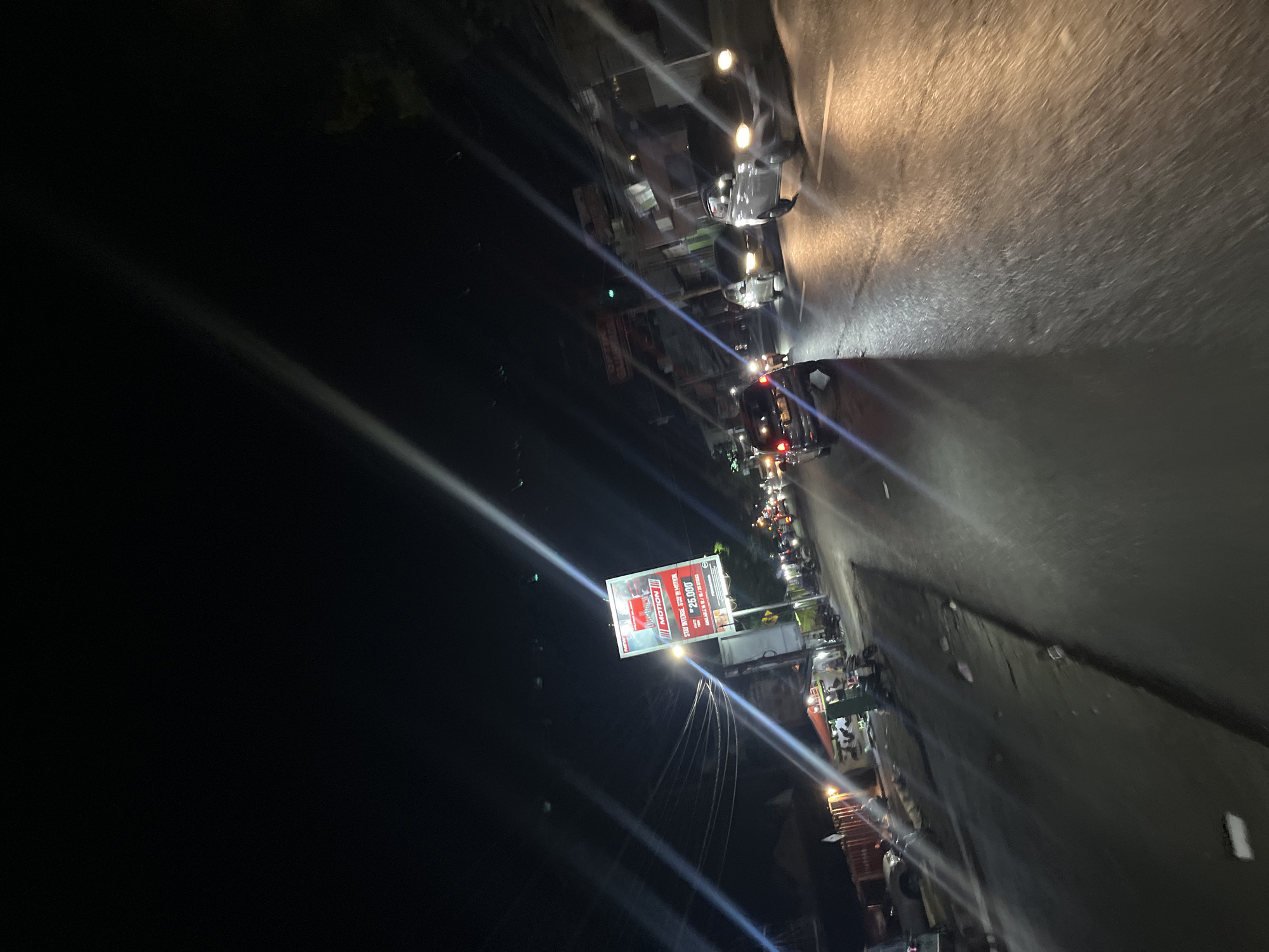This image captures a nighttime street scene in Kalinyamatan, Indonesia. The perspective is from a moving vehicle, looking forward and slightly upwards, with the camera tilted. The street is wet and reflects the headlights of oncoming traffic and streetlights, creating streaks of light across the asphalt.
Several cars and motorcycles are visible on the road, navigating through the night. Some cars have their headlights on, illuminating the path ahead. Motorcycles are parked on the side of the road and some are in motion.
On the left side of the street, there are buildings and shops. One prominent feature is a large billboard with red and white lettering, advertising "MOTION" and listing a price of "25.000". Power lines are strung overhead, crisscrossing the sky.
The sky is dark, indicating it is nighttime. The overall atmosphere is that of a typical urban street during evening hours, with activity and illuminated signs.