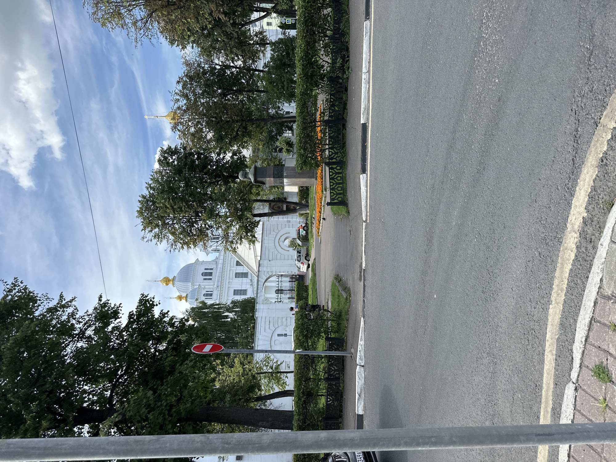 A sunlit, exterior medium shot captures a street scene in front of a white building with gold-domed roofs, likely a church or monastery, in Putyatino (Semivragovskaya S/A), Russia. The building is partially obscured by lush green trees. In the foreground, a gray asphalt road runs horizontally across the bottom of the frame. To the left, a portion of a silver car is visible. A gray metal pole stands prominently on the left side of the image.
A red and white "No Entry" sign is mounted on a pole to the left of a manicured green hedge. Behind the hedge, a person is walking towards a gated entrance of the white building. Several cars are parked or driving near the entrance. A bust monument on a pedestal is situated in front of the building, surrounded by a flower bed with bright orange flowers. The sky is blue with scattered white clouds.