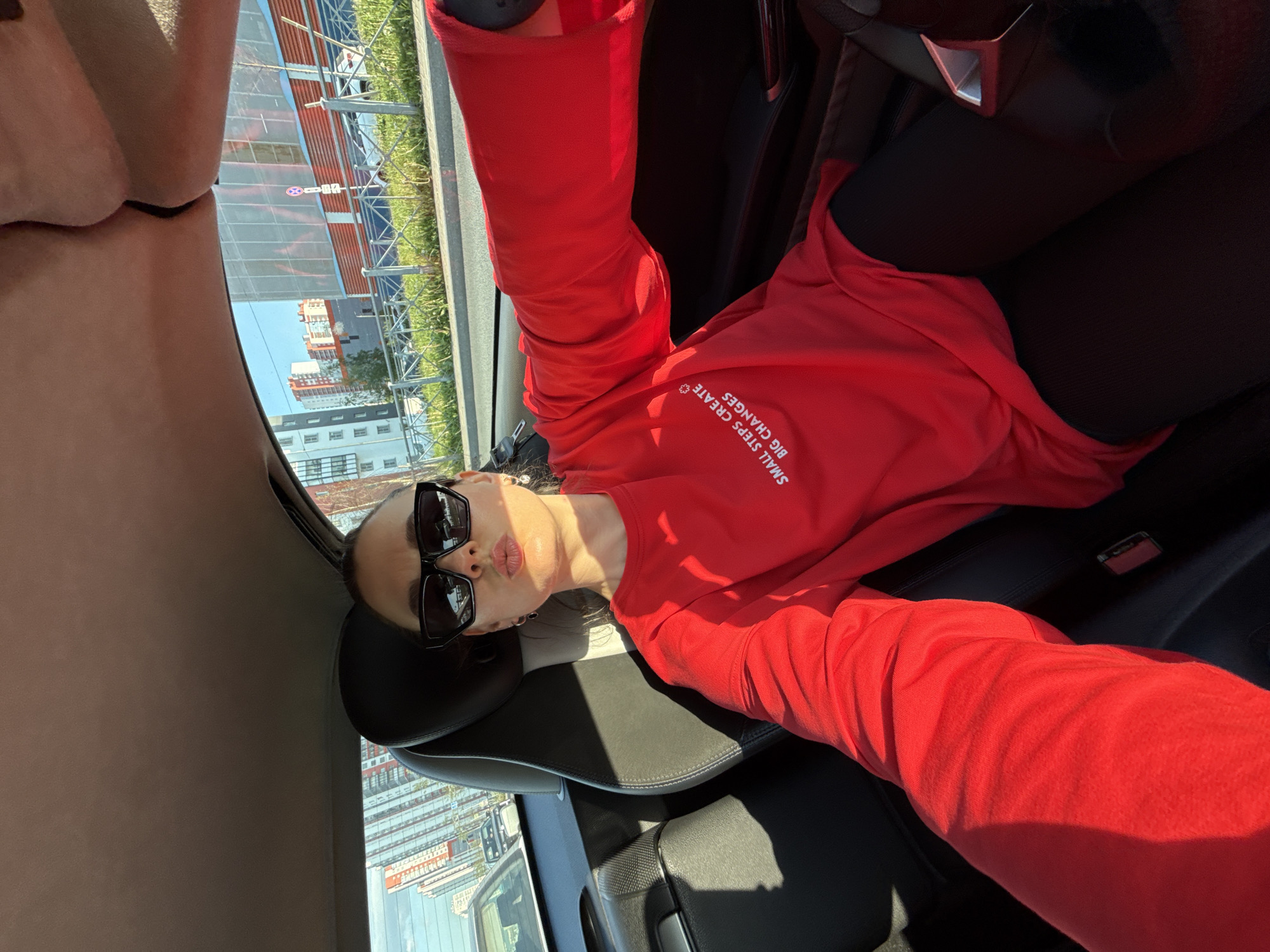 The image, rotated 90 degrees clockwise, captures a selfie of a person inside a car on a bright, sunny day in Saint Petersburg, Russia. The subject, likely female, is seated in what appears to be the driver's seat, looking towards the camera with pursed lips in a playful "kiss face" expression. She wears large, dark rectangular sunglasses and a bright red long-sleeved top. White text on her shirt reads, "SMALL STEPS CREATE BIG CHANGES". Her lower body is clad in black leggings.
The car's interior features dark upholstery, visible on the headrest and seats. Outside the car windows, a clear blue sky dominates the upper background. Below, a cityscape unfolds, showcasing modern buildings with numerous windows, suggesting an urban environment. In the mid-ground, there's what appears to be a construction site or industrial area with scaffolding, metal structures, and red-brown buildings. Green grass and a metal fence line the area closer to the car. A white circular sign with a red border and a diagonal line is also faintly visible in the background. Strong sunlight illuminates the scene, casting shadows within the car's cabin.