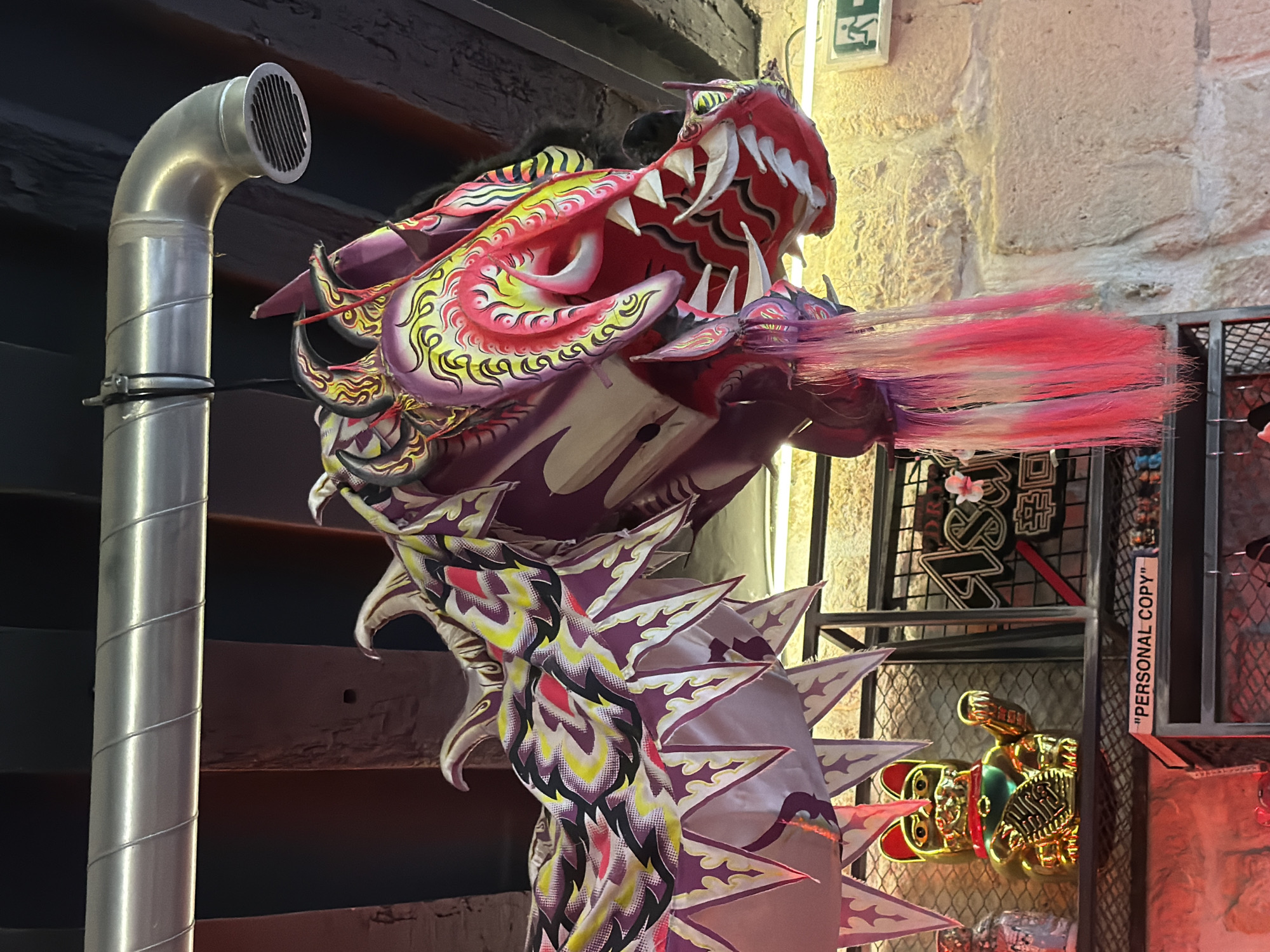 The image shows a colorful Chinese dragon decoration indoors, possibly a restaurant or store, with a red, pink, and purple color scheme. The dragon's head has prominent white teeth and long, pink whiskers. To the left of the dragon is a silver metal pipe. Behind the dragon is a textured light-colored wall and a metal grid shelf with a golden lucky cat figure and the words "PERSONAL COPY". The environment is dimly lit with a mix of natural and artificial lighting. This is located in the city of Paris, France.