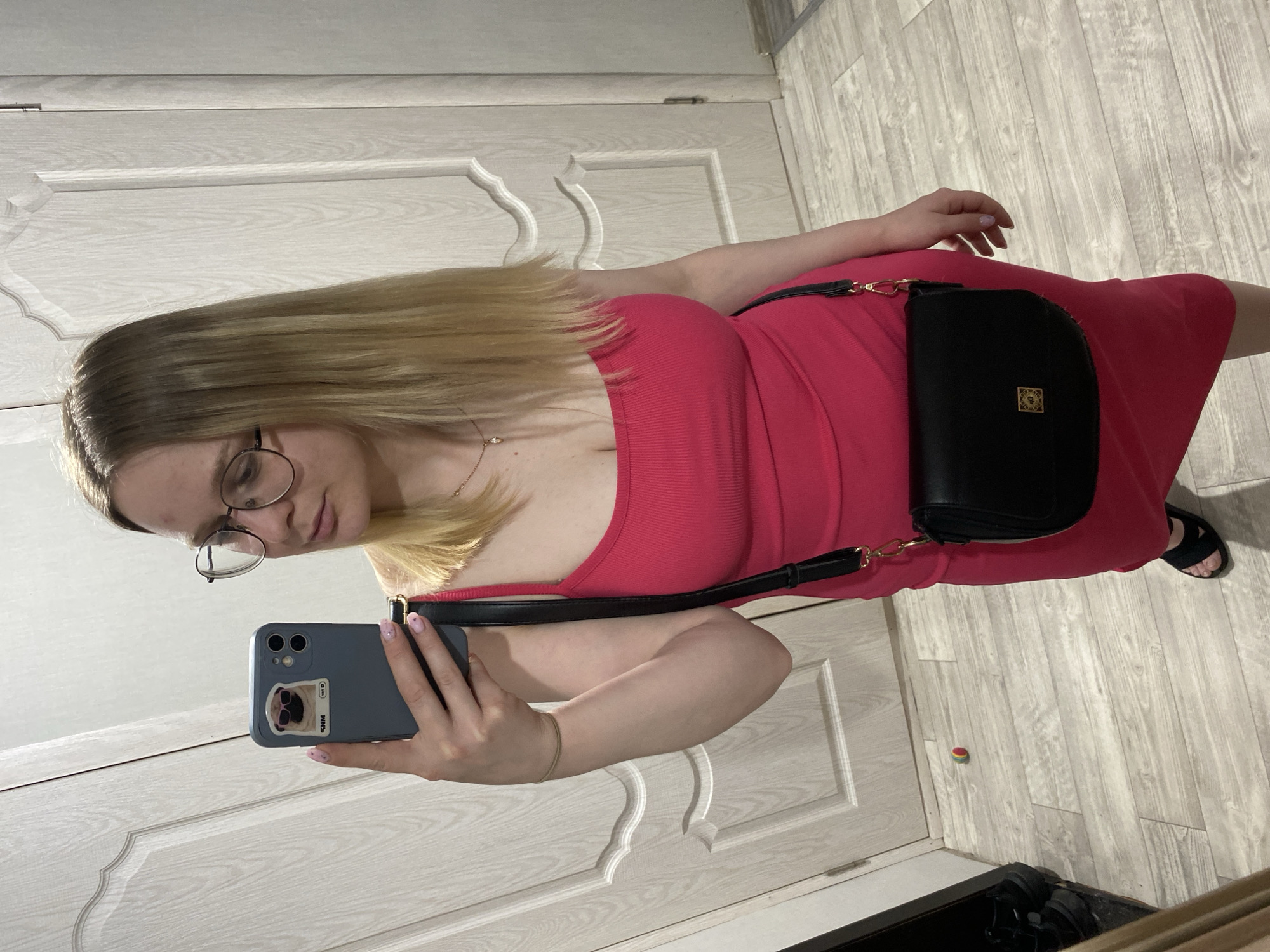 A woman stands indoors, in front of a closed white door with decorative carvings. She is taking a selfie with her phone, which has a light blue case and a dog sticker on it. The woman has blonde hair, wears glasses, and a pink dress. A black purse with a strap hangs over her shoulder. The floor is a light-colored wood-grain. The woman's feet are visible, she wears black sandals. The location is Chaikovsky, Russia.