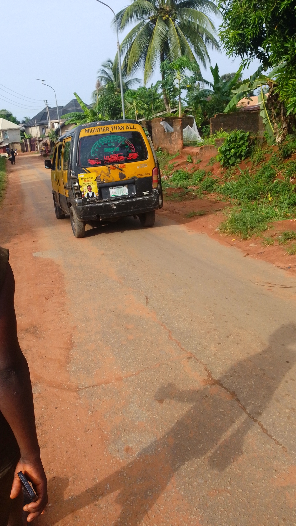 The image shows a yellow and black minivan with the text "MIGHTIER THAN ALL" on the back, driving on a partially paved road in Enugwu-Ukwu, Nigeria. The vehicle has a Nigerian license plate. The surrounding environment includes red dirt embankments with vegetation, palm trees, and residential buildings. A person holding a cell phone is visible in the lower left corner of the frame, and their shadow stretches across the road. Another person is walking along the side of the road in the distance.