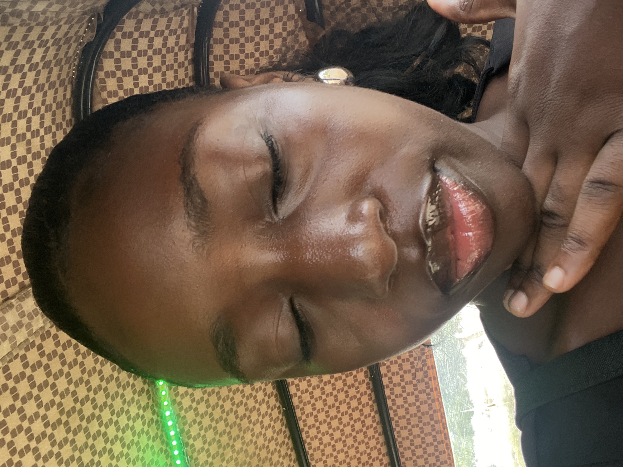 An African woman is lying down with her eyes closed. She is wearing black clothing. There is a hand near her face, and she has an earring in her left ear. The surface she is lying on has a checkered pattern. Green LED lights are visible. The photo does not provide enough information to determine that the location is Abuja, Nigeria.
