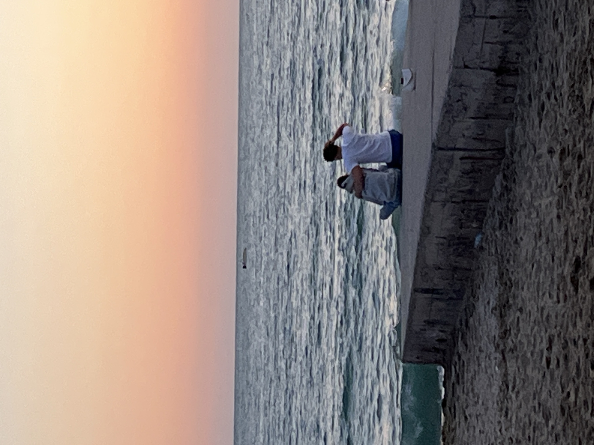 Two individuals are seated on a stone wall adjacent to a body of water. The person on the left is wearing a white shirt and dark pants, with their right hand raised towards their head. To their right, a person is leaning back, wearing a grey jacket. The water appears to be an ocean, with waves lapping against the wall. The sky in the background is a pale orange or pink color. The rough stone wall is adjacent to the water. Given the mention of Alanya, Turkey, it's plausible this scene is taking place on the coastline or a pier in that city.