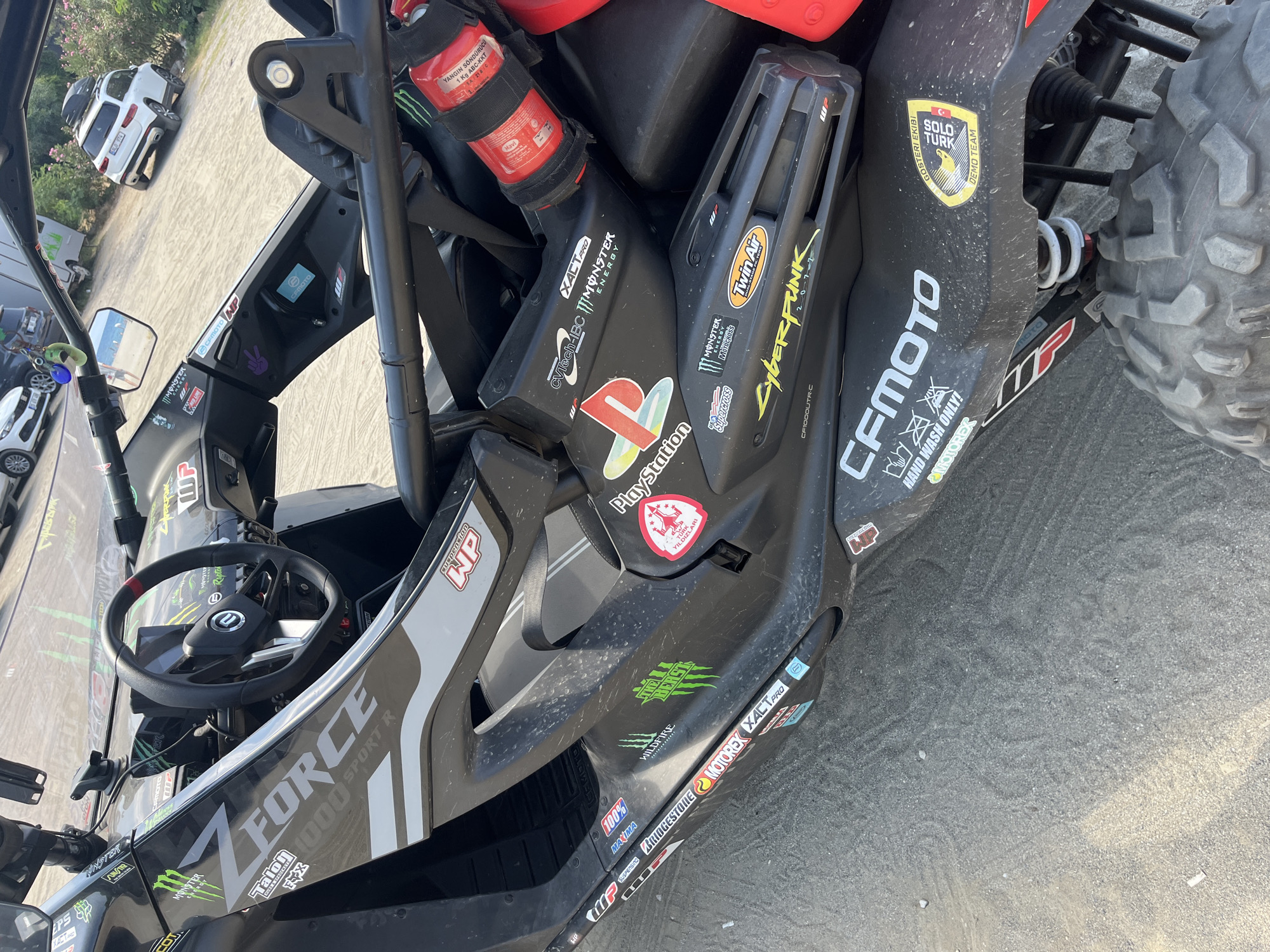 A detailed, forensic, matter-of-fact description of the scene:
The image captures the interior and exterior of a black all-terrain vehicle (ATV), likely a CFMOTO ZFORCE 1000 SPORT R, parked on a sandy, gravelly surface. The ATV is heavily adorned with various stickers, including logos for Monster Energy, PlayStation, Cyberpunk 2077, Wildfire, Motorex, Bridgestone, and others. A red fire extinguisher is mounted on the roll cage. The steering wheel and dashboard are visible, with a reflection of the sky and a beach in the side mirror.
In the background, a white SUV with a roof rack is parked on a dirt road, with a body of water and trees visible beyond. The setting appears to be a parking area near a beach, consistent with the city of Alanya, Turkey, based on the "GÖSTERİ EKİBİ SOLO TÜRK DEMO TEAM" sticker, which indicates a Turkish demonstration team. No people are visible in the frame. The environment suggests an outdoor recreational or event location.