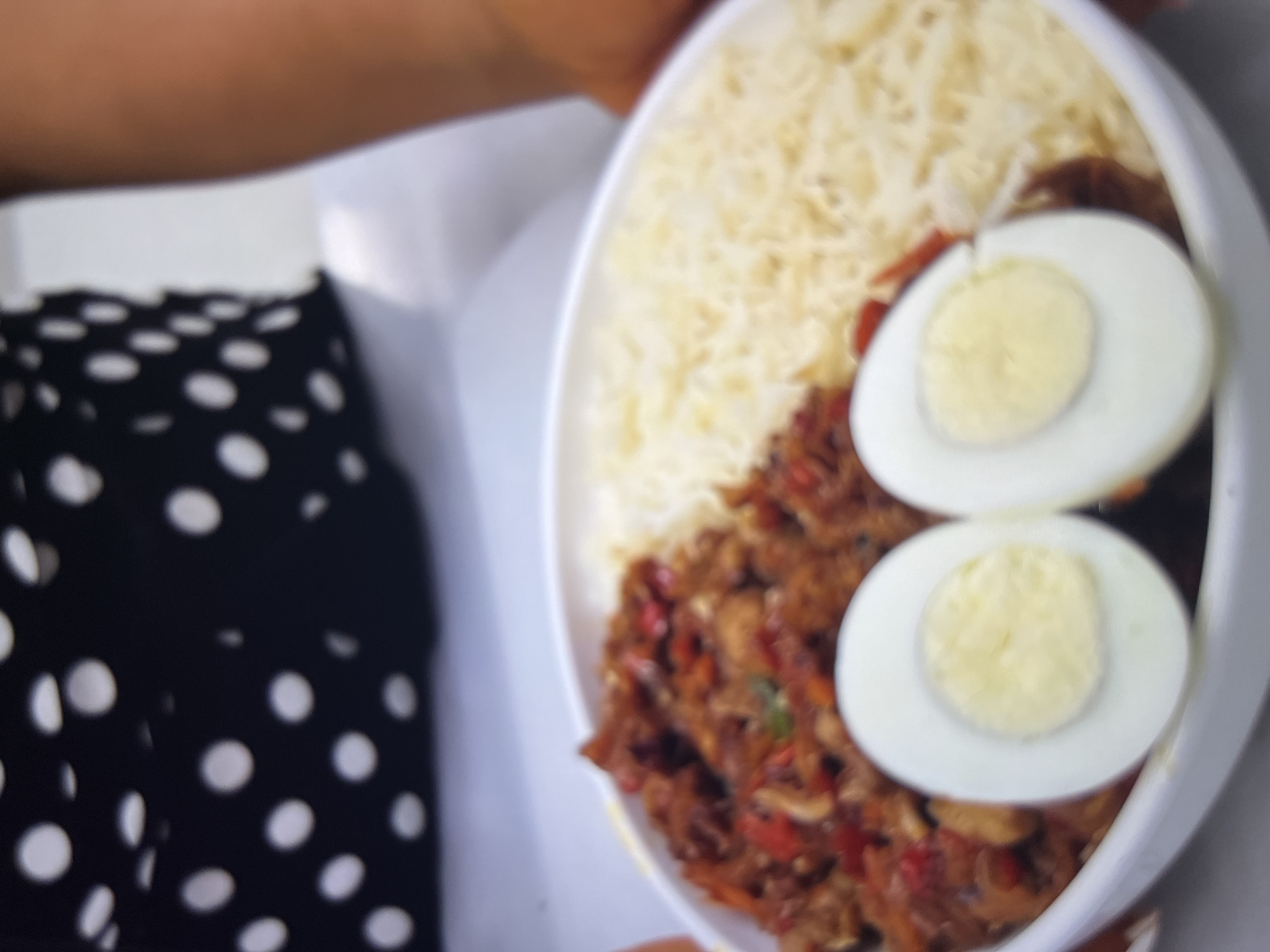 A close-up shot shows a white bowl of food. The bowl is divided into two sections, with white rice filling the right half and a dark, reddish-brown stew filling the left half. The stew appears to contain small pieces of meat and vegetables. Two halves of a hard-boiled egg are placed on top of the stew, with the yolks facing upwards. A person's arms and torso, wearing a black and white polka-dotted top, are visible in the background, holding the bowl. The lighting is soft and diffused, suggesting an indoor setting. There is no visible indication of the location being in Abuja, Nigeria.