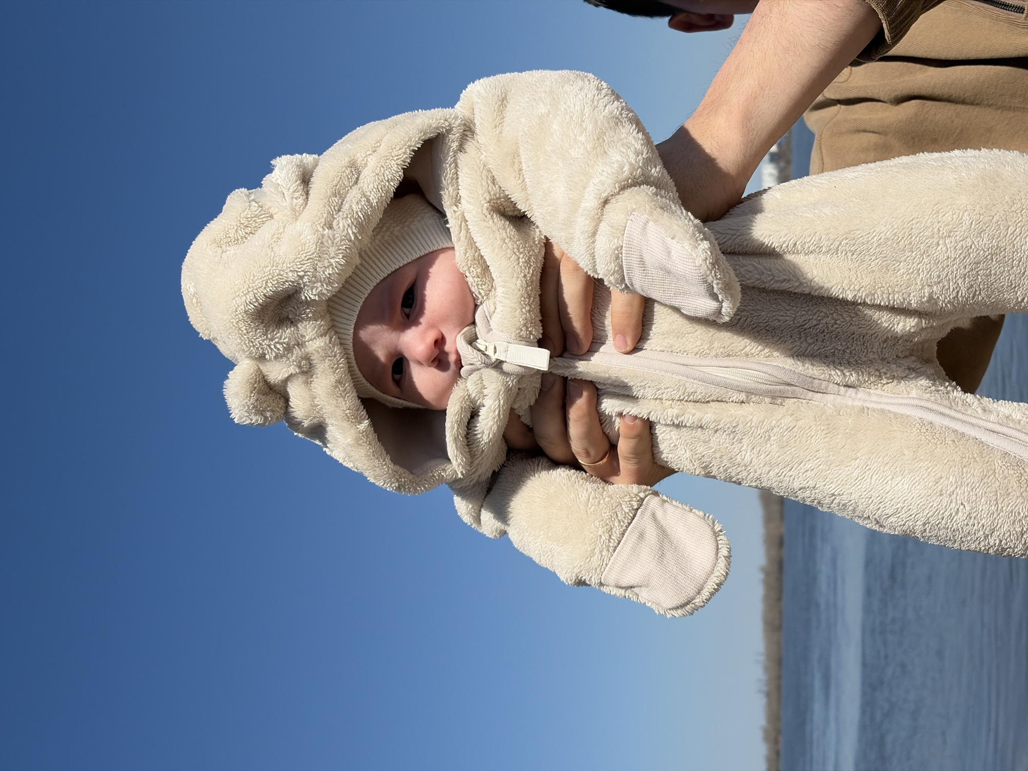 A baby wearing a fluffy, cream-colored hooded jumpsuit is being held up against a bright blue sky. The person holding the baby is wearing a brown top. The background reveals a sliver of water in the city of Noviy, Russia.