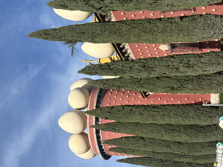 The image displays a unique architectural structure, likely a museum or gallery, set against a clear blue sky. The most striking features of the building are numerous large, egg-shaped sculptures, light beige in color, perched prominently along the rooftop edges. These eggs are rounded and smooth, varying slightly in size and placement.
The main facade of the building is a deep terracotta red, extensively adorned with a repetitive pattern of small, golden, bread-like shapes, resembling baguettes or loaves, which are affixed to the surface. Towards the top of the building, beneath the eggs, there are also some golden, statue-like figures, including what appears to be a classical figure with arms outstretched, adding to the ornate and surreal aesthetic.
In the foreground, several tall, slender, dark green coniferous trees, resembling cypress trees, stand vertically, partially obscuring the building's facade and some of the egg sculptures. These trees are uniformly shaped and create a strong vertical element in the composition.
The sky is a vibrant blue with a few faint, wispy white clouds visible, indicating a clear, sunny day. In the lower right portion of the image (when oriented correctly), some additional architectural elements or structures are visible, including what might be scaffolding or metal framework and possibly some equipment or a ladder, suggesting an active area or ongoing work, or simply part of the complex’s design. No people or visible text are present in the image. The overall impression is one of distinctive, surreal architecture under bright daylight.