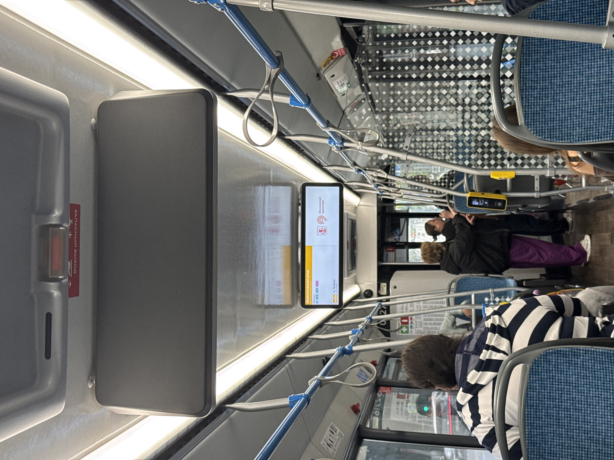 The interior of a public transport vehicle, likely a bus, in Moscow, Russia, is depicted. The perspective is from a seated passenger looking forward. The vehicle is equipped with blue and silver handrails and straps for passengers. A digital display screen is mounted above the aisle, showing route information including "Шереметьевская улица" (Sheremetyevskaya Street) and route numbers "512" and "519". Below this, another route "M35a" is listed with "м. Трубная" (Trubnaya metro station). A logo for "Московский транспорт" (Moscow Transport) is visible on the screen.
Several passengers are present. In the foreground, a person wearing a black and white horizontally striped hooded jacket is seated. Further down the aisle, two individuals are standing near the front. One, in the middle ground, wears a black jacket and purple pants, and the other, to their right, wears a dark jacket. The interior features a patterned divider or wall with a repeating diamond motif. The lighting appears to be from overhead fluorescent fixtures. The scene captures a typical moment of transit within the city's public transportation system.