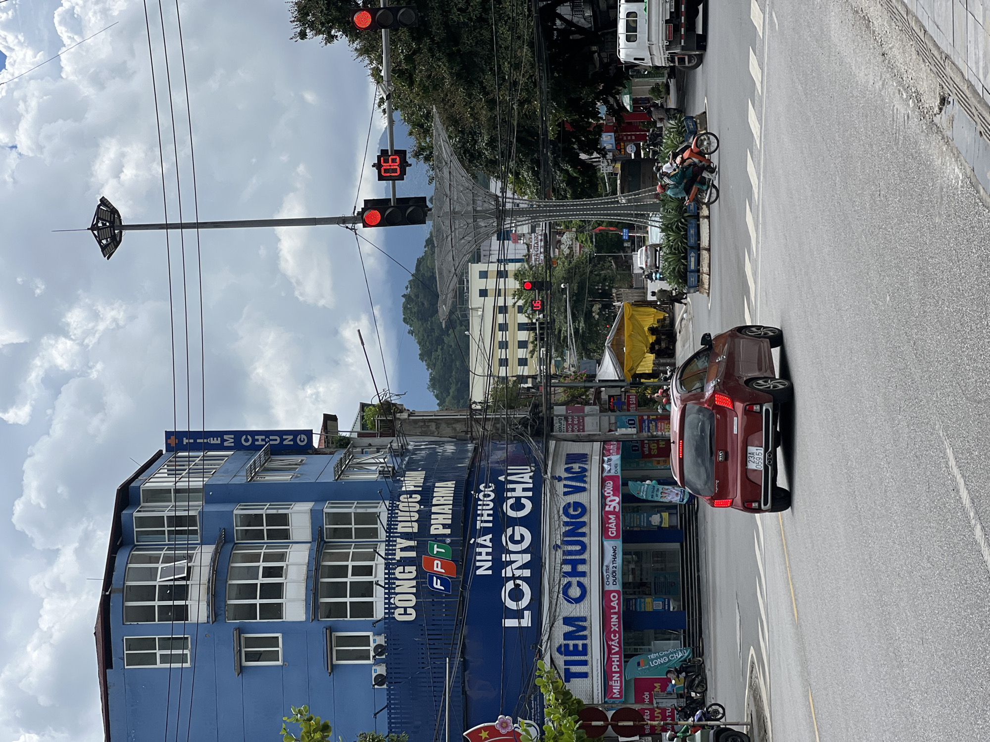 This is a street-level, eye-level shot of an intersection in Hà Giang 2 Ward, Vietnam. The scene is dominated by a large, bright blue building on the left, which has multiple floors and many windows. A vertical blue sign with white text reading "TIEM CHUNG" is affixed to the building's facade. Below this, larger white text reads "NHA THUOC LONG CHAU." Further down, another blue sign with white text reads "TIEM CHUNG VAC."
On the right side of the image, a tall street light stands prominently, supporting traffic signals and a digital display showing "06." There are also trees and other buildings visible in the background. The street is paved with asphalt, and there are crosswalk markings.
In the foreground, a red Kia car is driving away from the viewer, its license plate reading "23A 059.51." Several motorcycles are also present, some parked and others in motion. People are visible on the motorcycles and walking on the sidewalk. A white truck is parked on the far right. The sky is partly cloudy. The overall atmosphere suggests a busy urban environment.