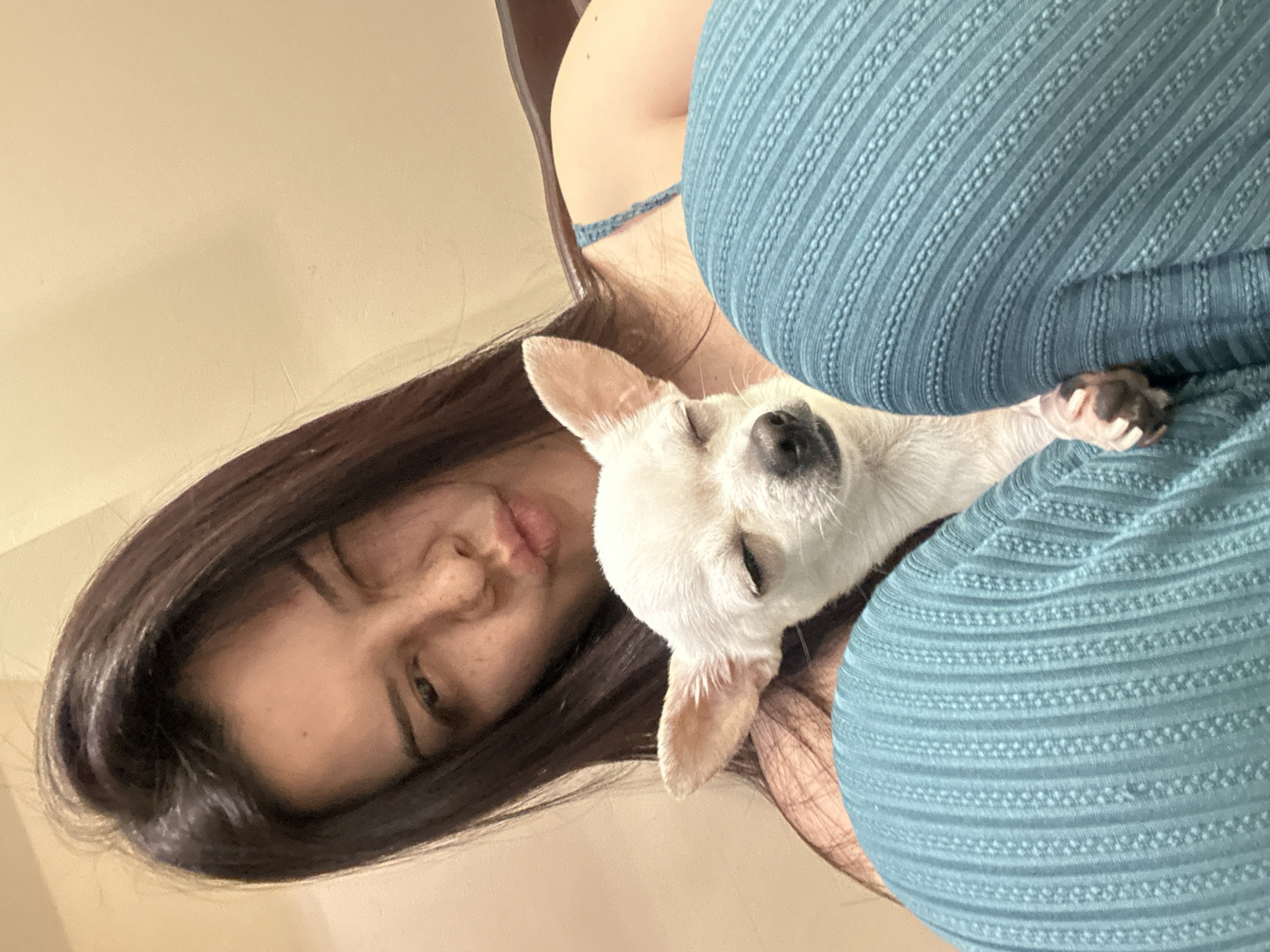 A selfie-style photograph shows a woman with dark brown hair, fair skin, and freckles, looking at the camera with one eye partially closed and her lips slightly pursed. A small, white Chihuahua with closed eyes is nestled against her, seemingly asleep. The woman is wearing a teal, textured top, and a small portion of a wooden chair back is visible in the upper left. The background is a plain, light beige wall. There is no information in the image to suggest a location in the city of Sliven, Bulgaria.