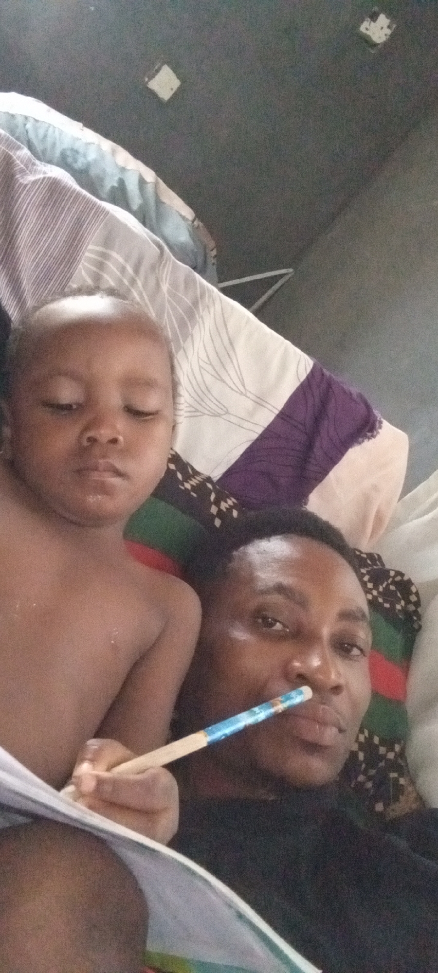 A man and a young child are lying down, presumably on a bed. The man is holding a pen or pencil near his mouth. The background includes pillows and a wall with some rectangular patches or fixtures. The image was taken in Adankolo, Nigeria.
