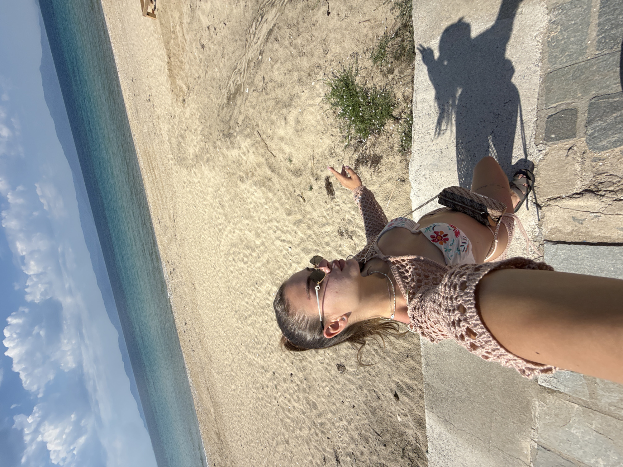 A woman in a bikini and crocheted top is seen on a sandy beach next to a sea. She is sitting on a stone structure, likely a low wall or steps, and facing the beach and sea. She is wearing sunglasses. The location is Unknown, Unknown. The sea is blue, and the sky is partly cloudy. The beach is sandy with some vegetation.