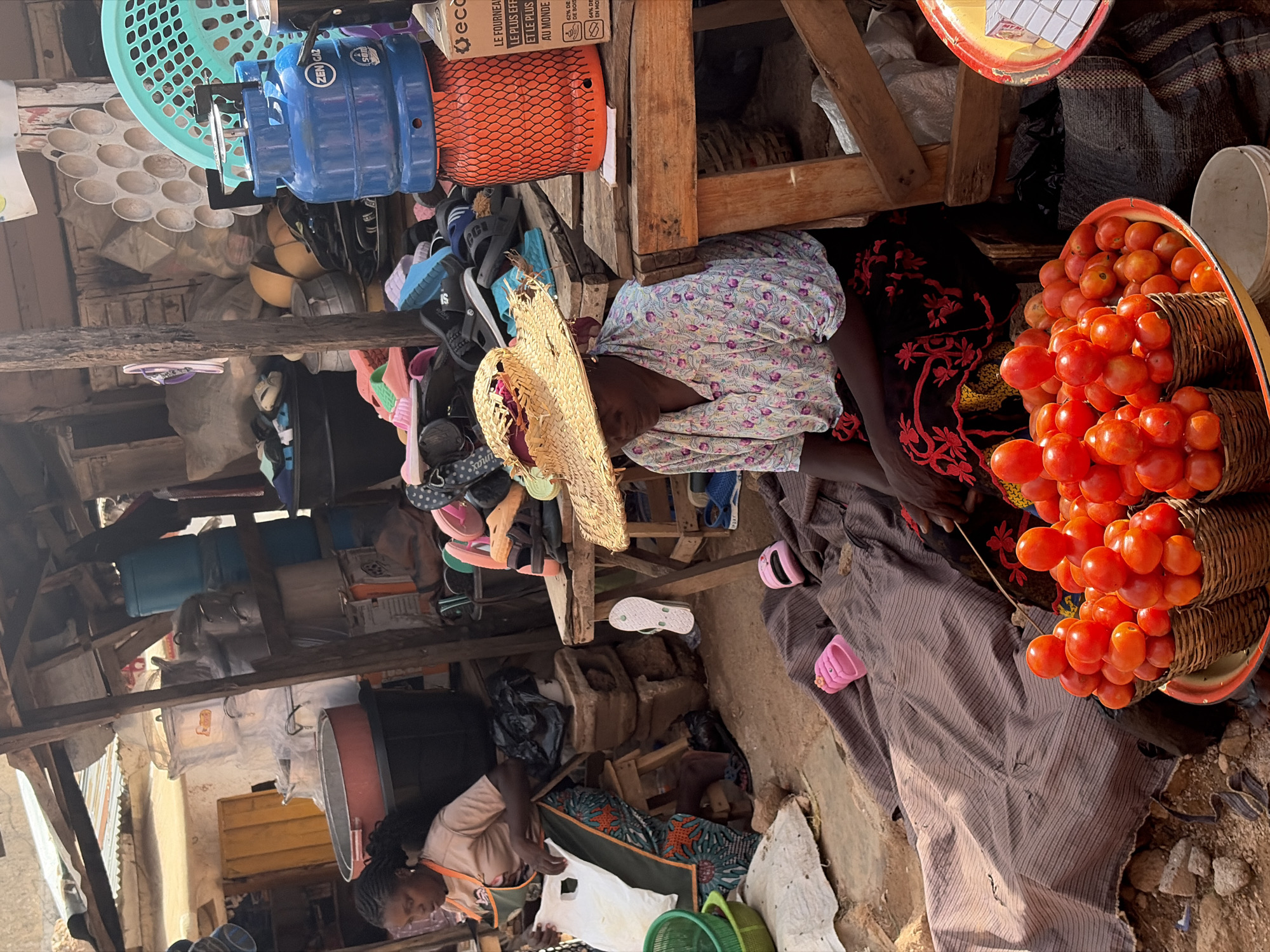In Jos, Nigeria, a woman is seated on a stool at a market stall, wearing a patterned shirt and a dark skirt. She has a straw hat on her head. In front of her are baskets filled with tomatoes, and a large tray also containing tomatoes. To her left, another woman is seated, wearing an apron over her clothes. Behind the seated women, the market stall is filled with various items including plastic containers, buckets, cooking pots, and a blue gas cylinder. Several pairs of sandals and flip-flops are visible. A large grey fabric is draped on the ground in the foreground, with pink children's slippers on top of it. The ground is dirt and the market appears to be outdoors, with wooden structures and corrugated metal roofing.