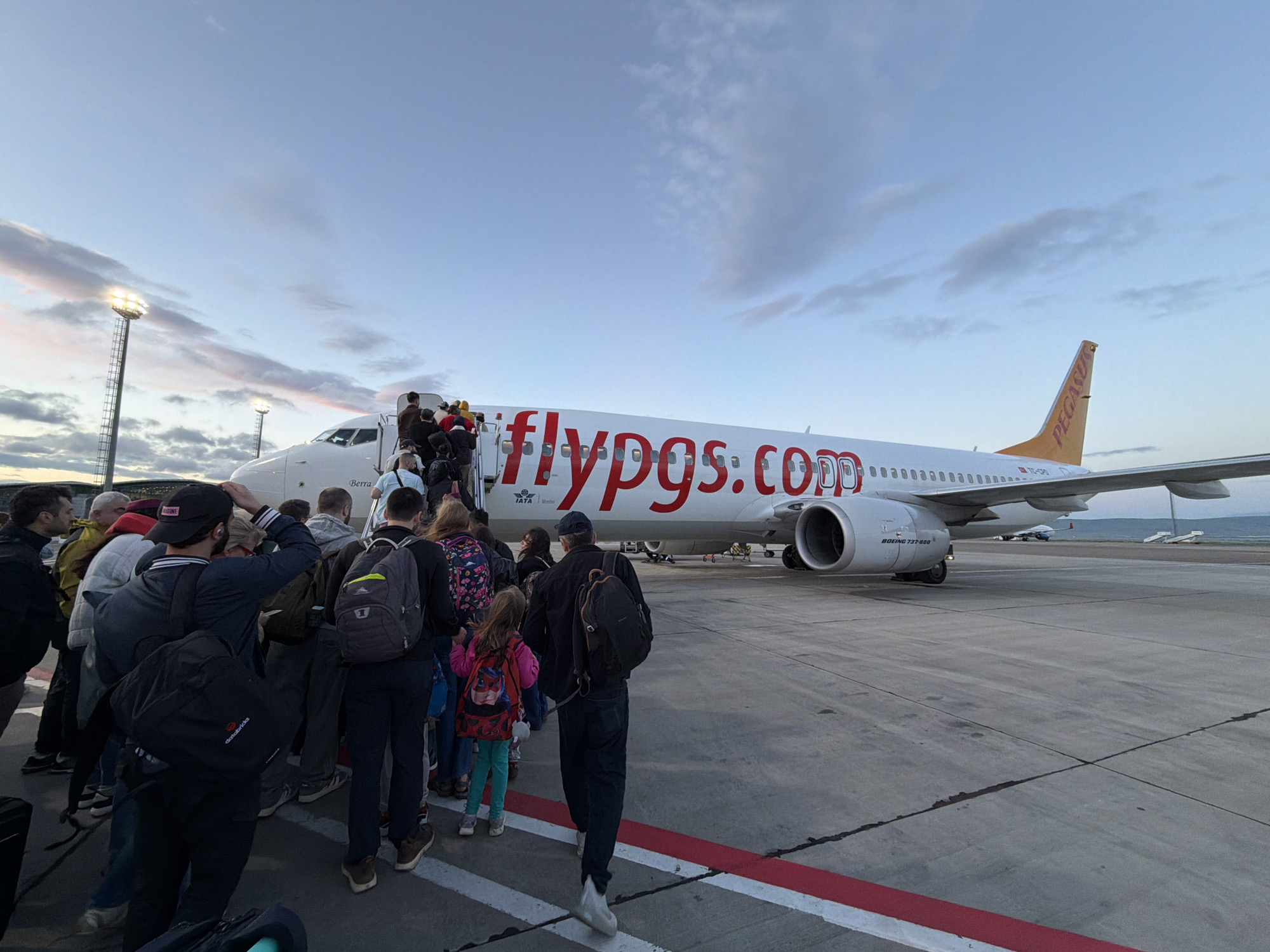 Passengers are boarding a white Pegasus Airlines Boeing 737-800 aircraft. The aircraft's registration is TC-CPR. The tail is orange with a Pegasus logo. People are gathered near the stairs leading to the plane’s entrance. Many have backpacks. In the background, the sky is blue with some clouds. The setting appears to be an airport tarmac. There is airport equipment and a second plane visible in the background. The lighting suggests it's either dawn or dusk. While the city cannot be definitively verified as Tbilisi, the image matches the general appearance of boarding at Tbilisi International Airport based on available online images.