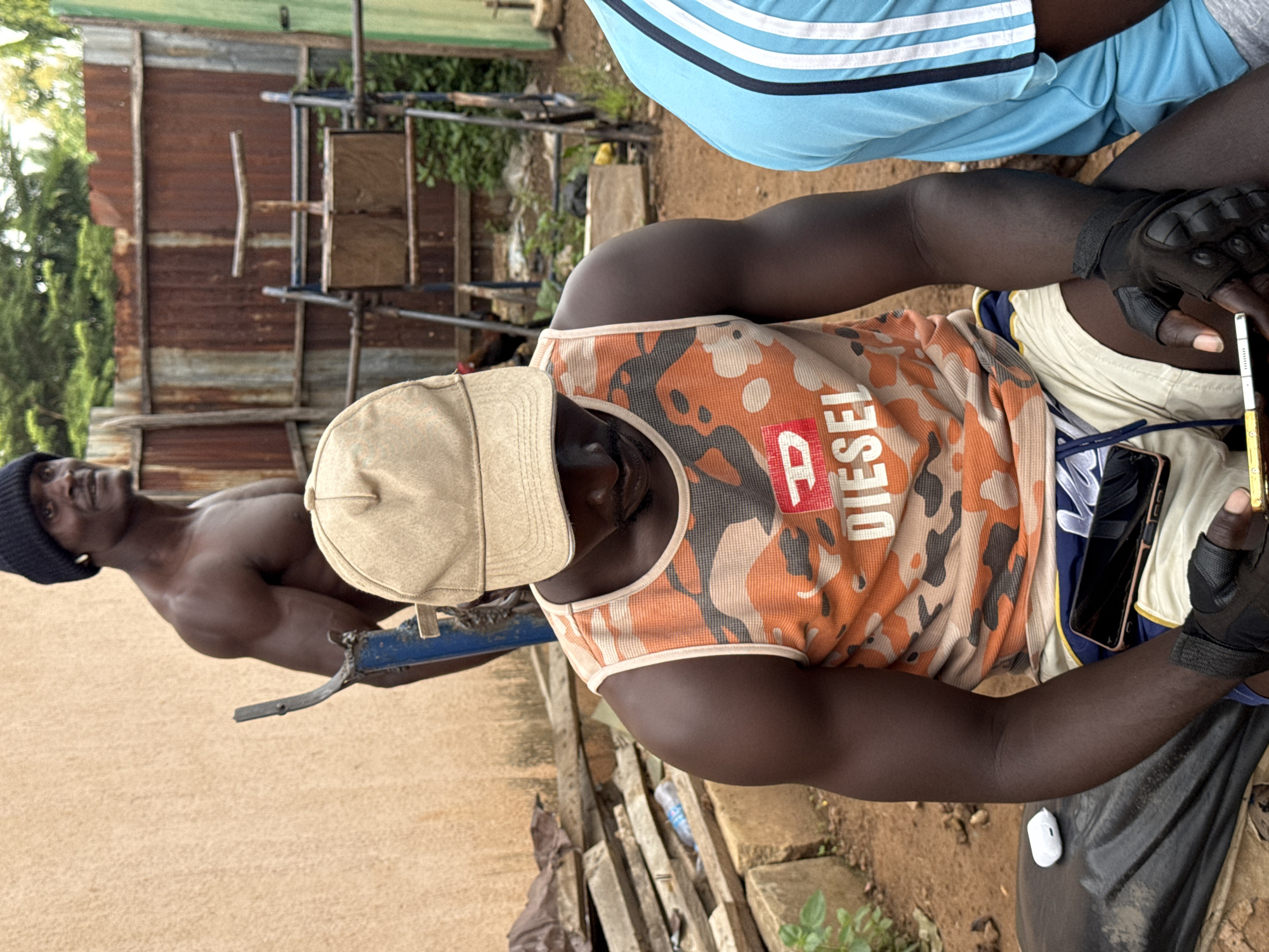 In Akwanga, Nigeria, a man wearing a black beanie and a black tank top is seen from the waist up, flexing his arm. He is standing behind another man who is seated and wearing a tan baseball cap and an orange and black camouflage tank top with "DIESEL" written on it in white letters. The seated man is looking down at a smartphone he is holding. To the right of the seated man, another person wearing a light blue shirt with white stripes on the sleeves is partially visible. The environment appears to be an outdoor gym or training area, with a corrugated metal fence in the background, along with gym equipment and stacks of wood. The ground is covered in dirt.