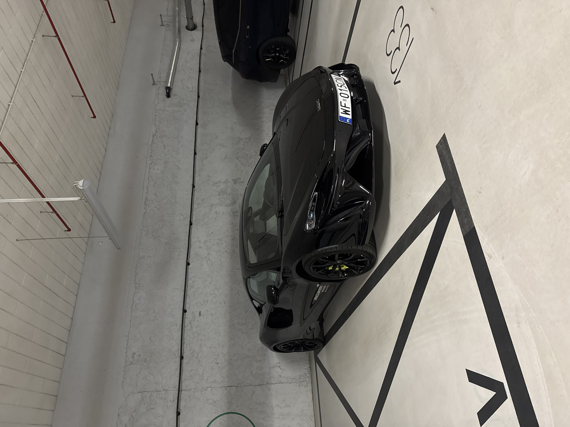 The image shows an indoor parking garage in Warsaw, Poland. A black McLaren sports car is parked in space 33. A second dark car is partially visible in the upper portion of the image. The McLaren has a license plate of "WF 015CM". The parking space is marked with black directional arrows. The garage has white walls and ceiling, with exposed pipes and conduit.